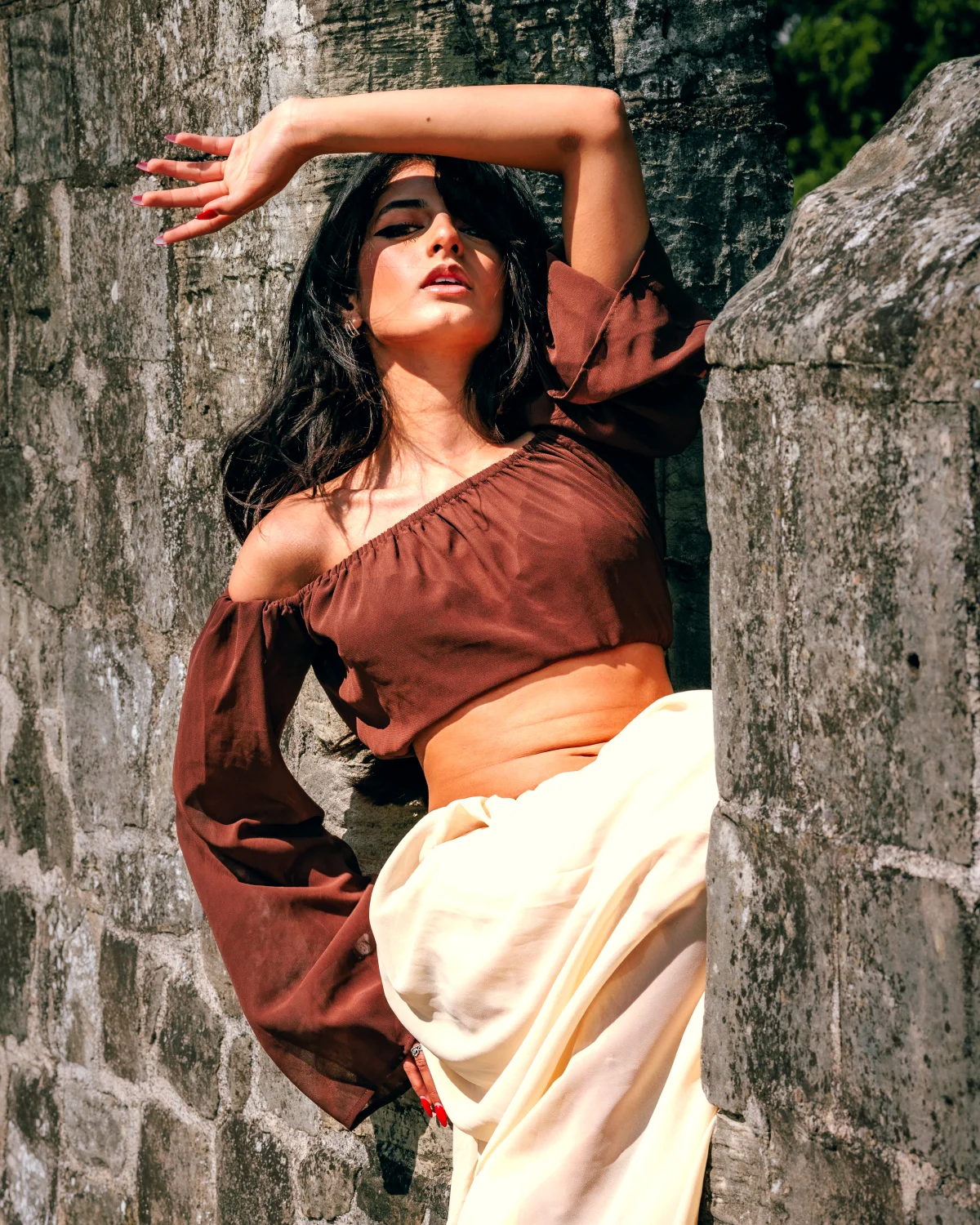 A woman in a brown off-the-shoulder crop top and cream skirt posing against a weathered stone wall.