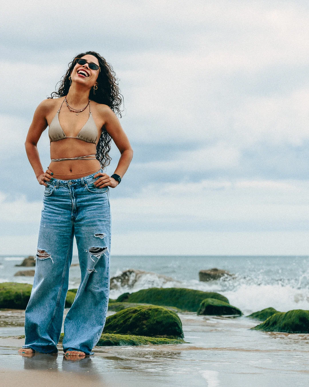 Smiling woman with curly hair in a bikini top and ripped blue jeans posing on a rocky beach.