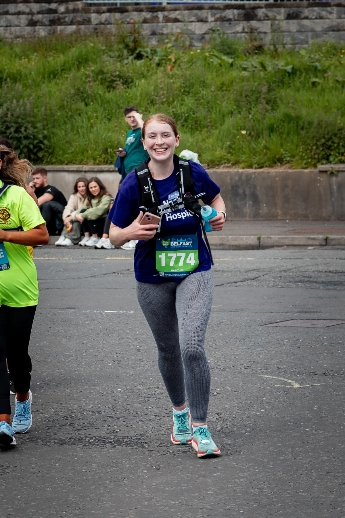 Smiling woman in a blue race shirt and grey leggings running during the Belfast marathon.