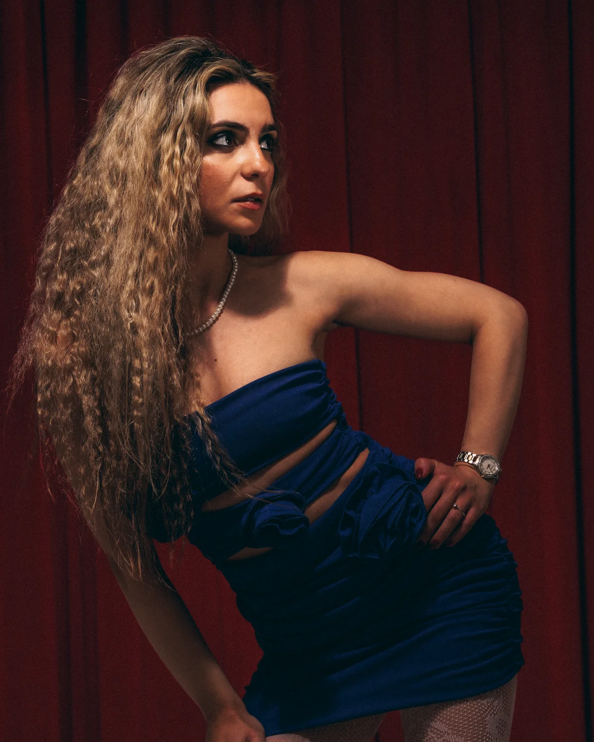 Woman with long curly blonde hair in a blue strapless dress posing against a red curtain backdrop.