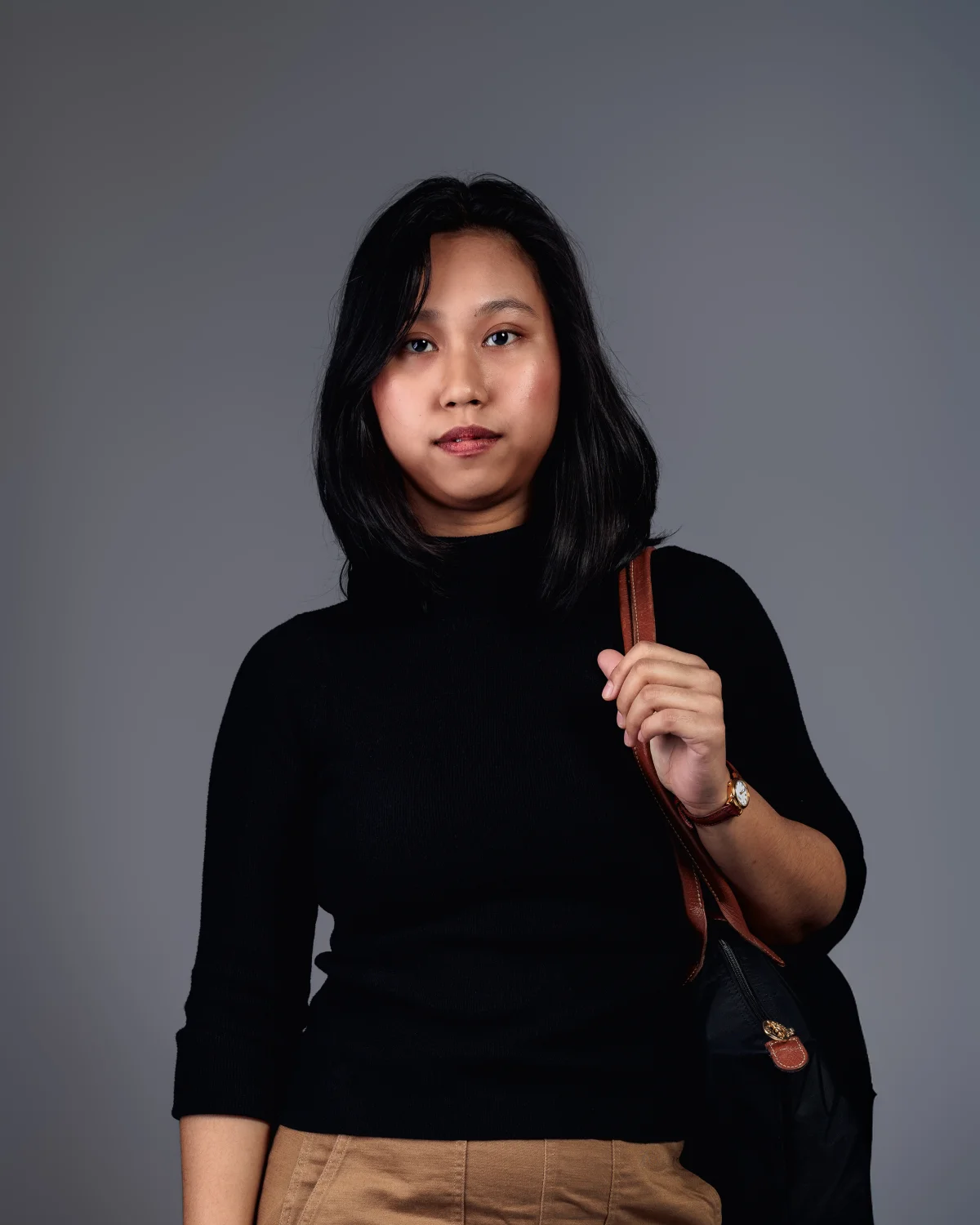 Studio portrait of a woman in a black turtleneck and tan trousers against a neutral grey background.
