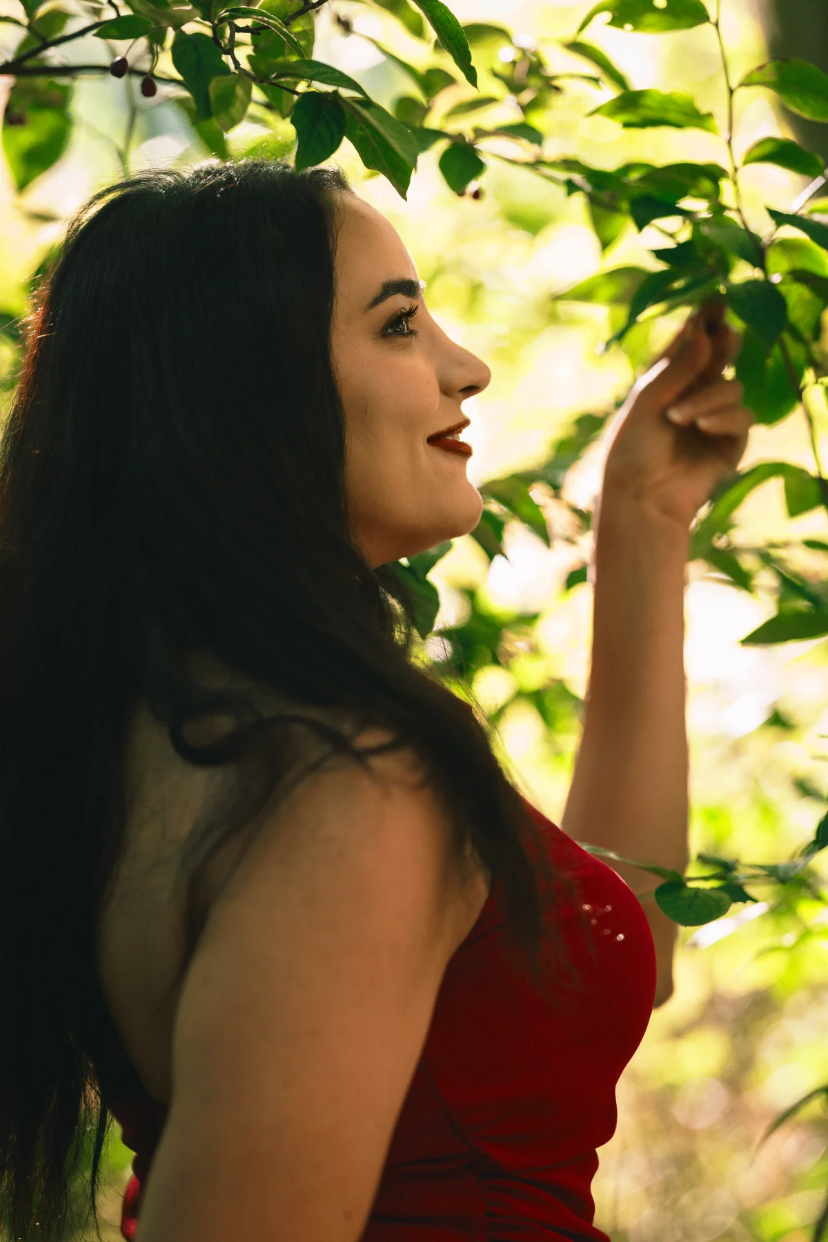 Side profile of a woman with long dark hair in a red dress among lush green leaves.