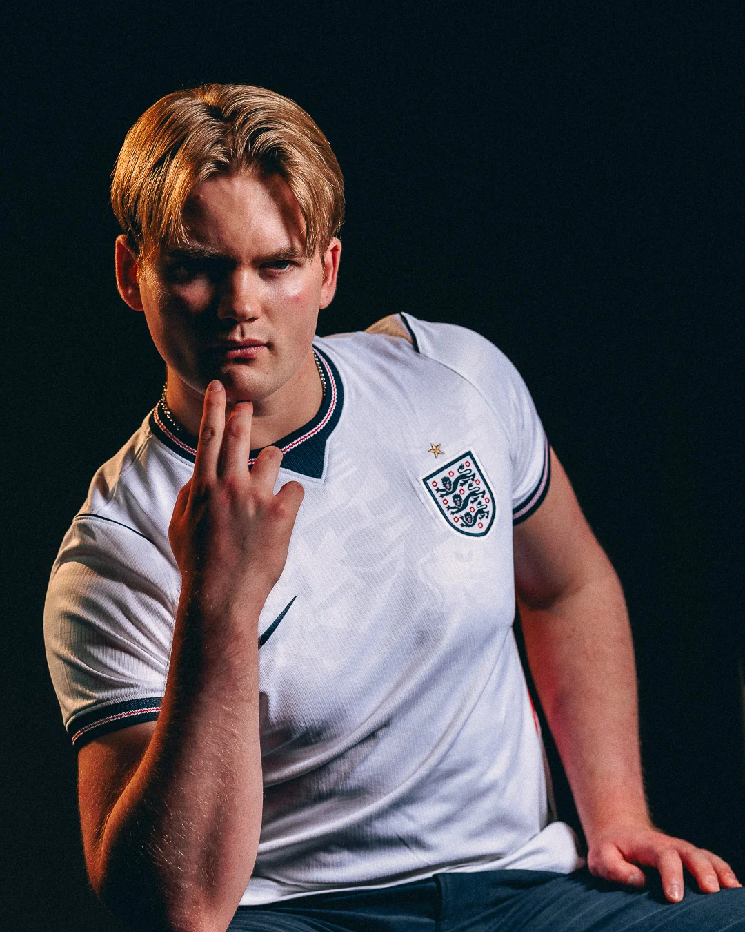 A moody studio portrait of a young man with blond hair wearing a white England football jersey.