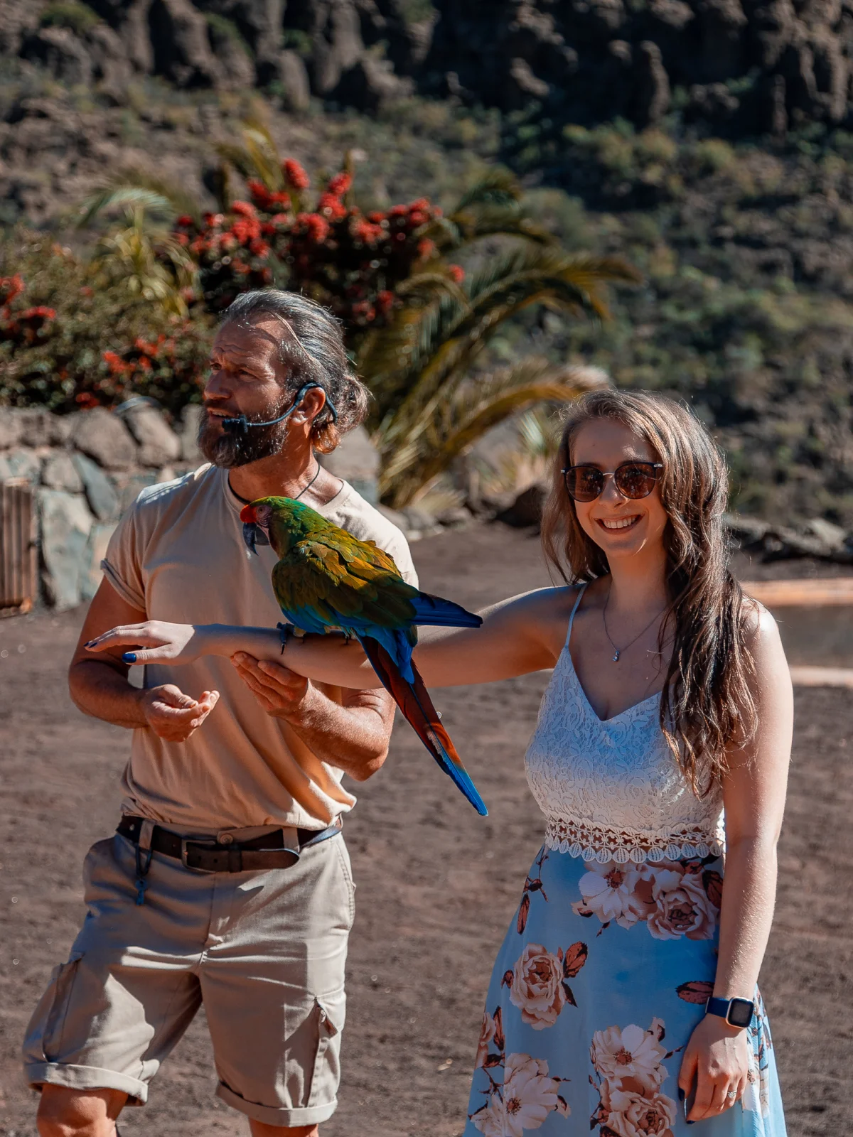A man and woman smiling while holding a colorful parrot in a rocky outdoor landscape.