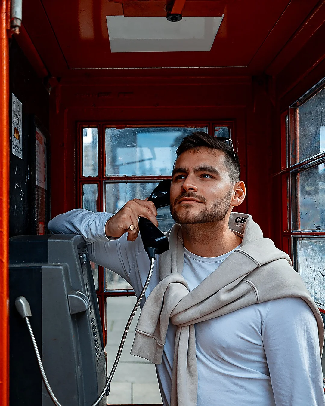 A man in a white shirt and beige sweater posing with a phone inside a red telephone booth.