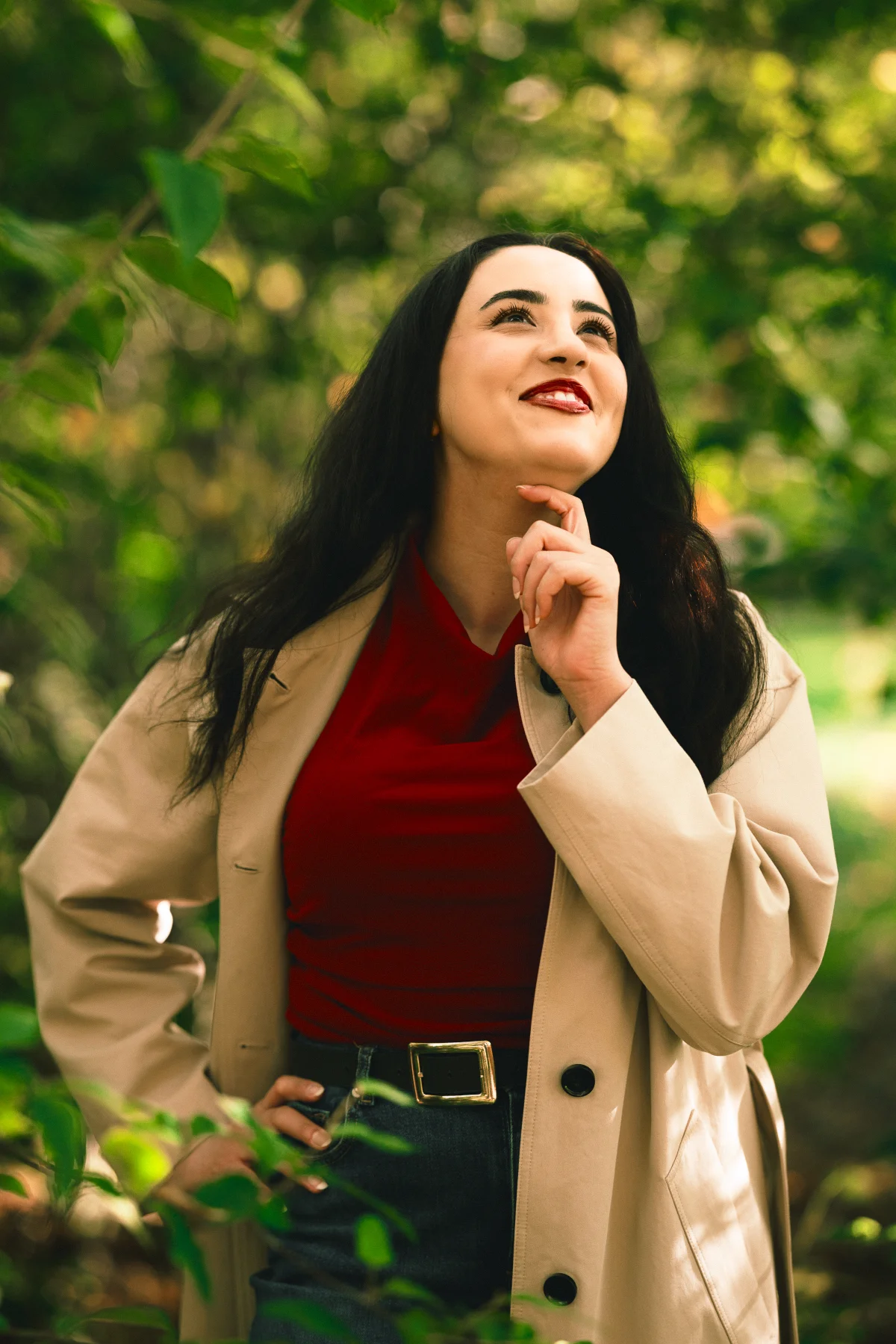 A young woman with long dark hair in a red top and beige coat, looking up thoughtfully in a green forest.