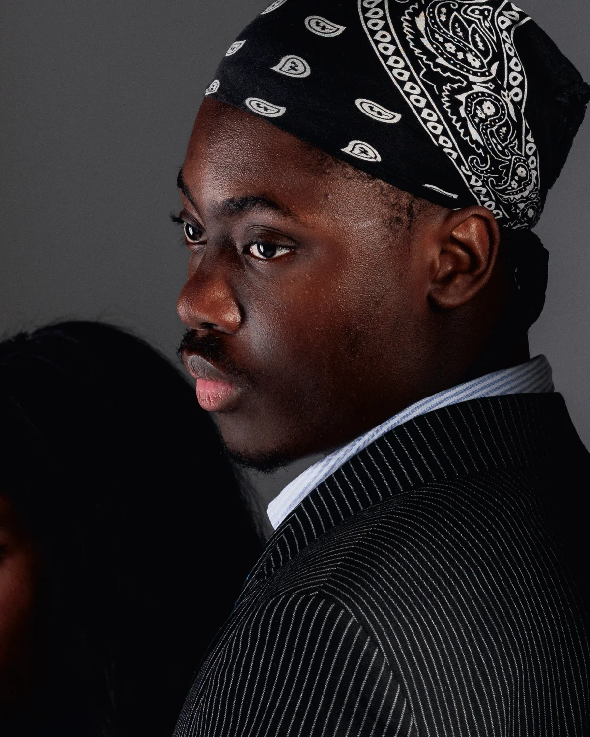 Side profile portrait of a Black man wearing a paisley bandana and pinstripe suit jacket against a grey background.