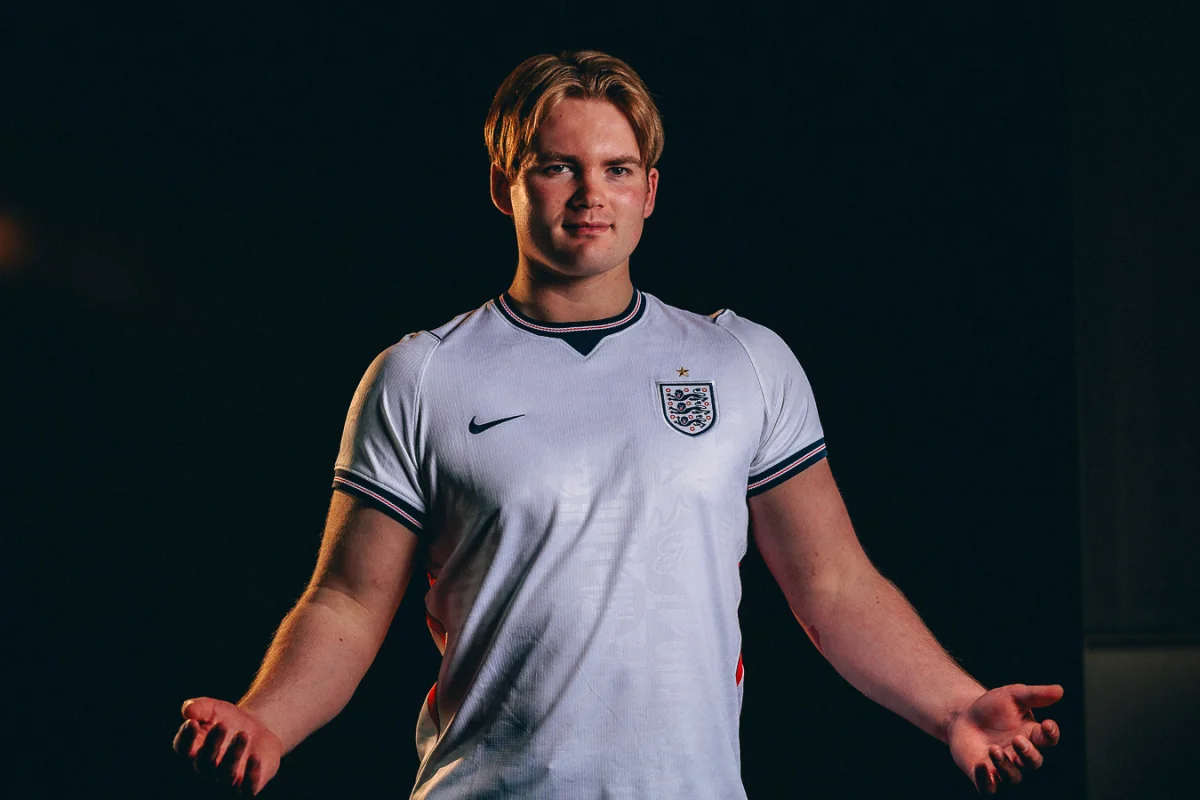 A young man with blond hair wearing an England football jersey posing against a dark studio background.