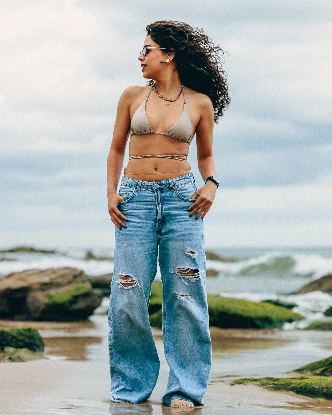 Woman with curly hair in a beige bikini top and ripped wide-leg jeans posing on a rocky beach.