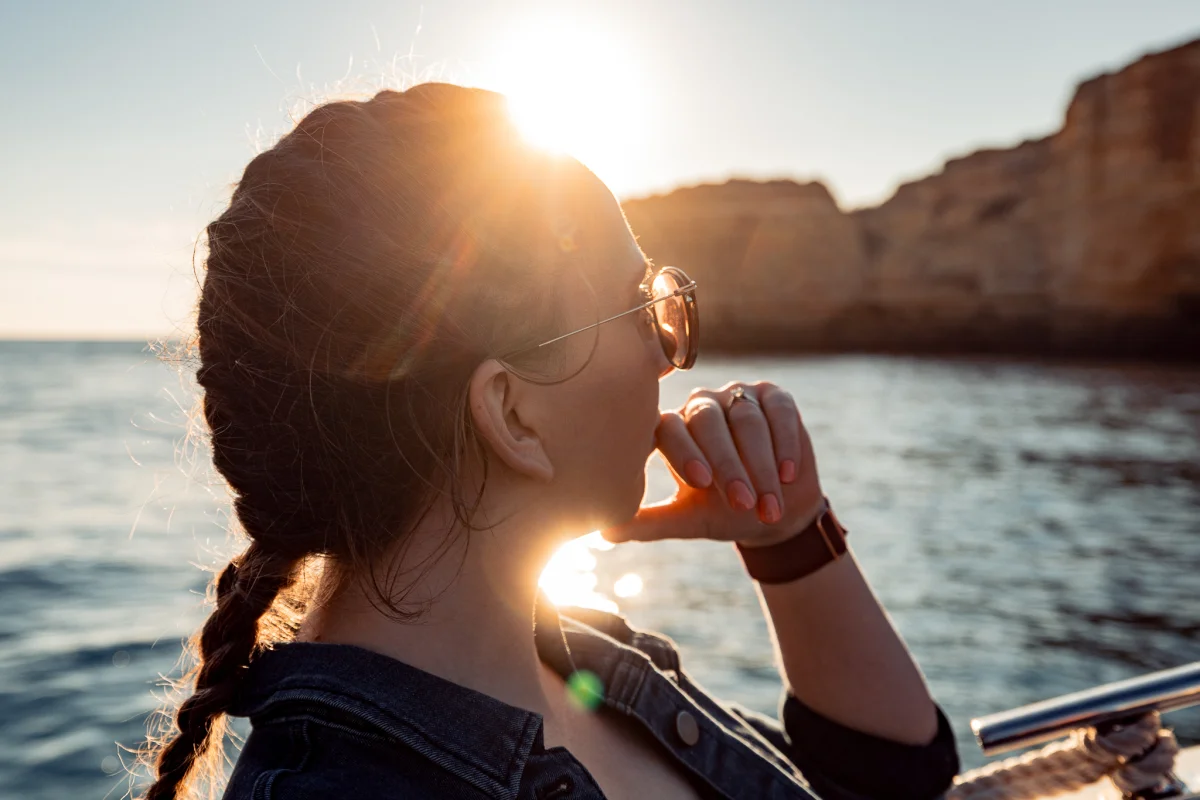 Profile view of a woman wearing sunglasses and a denim jacket, gazing at a sunset over ocean cliffs.