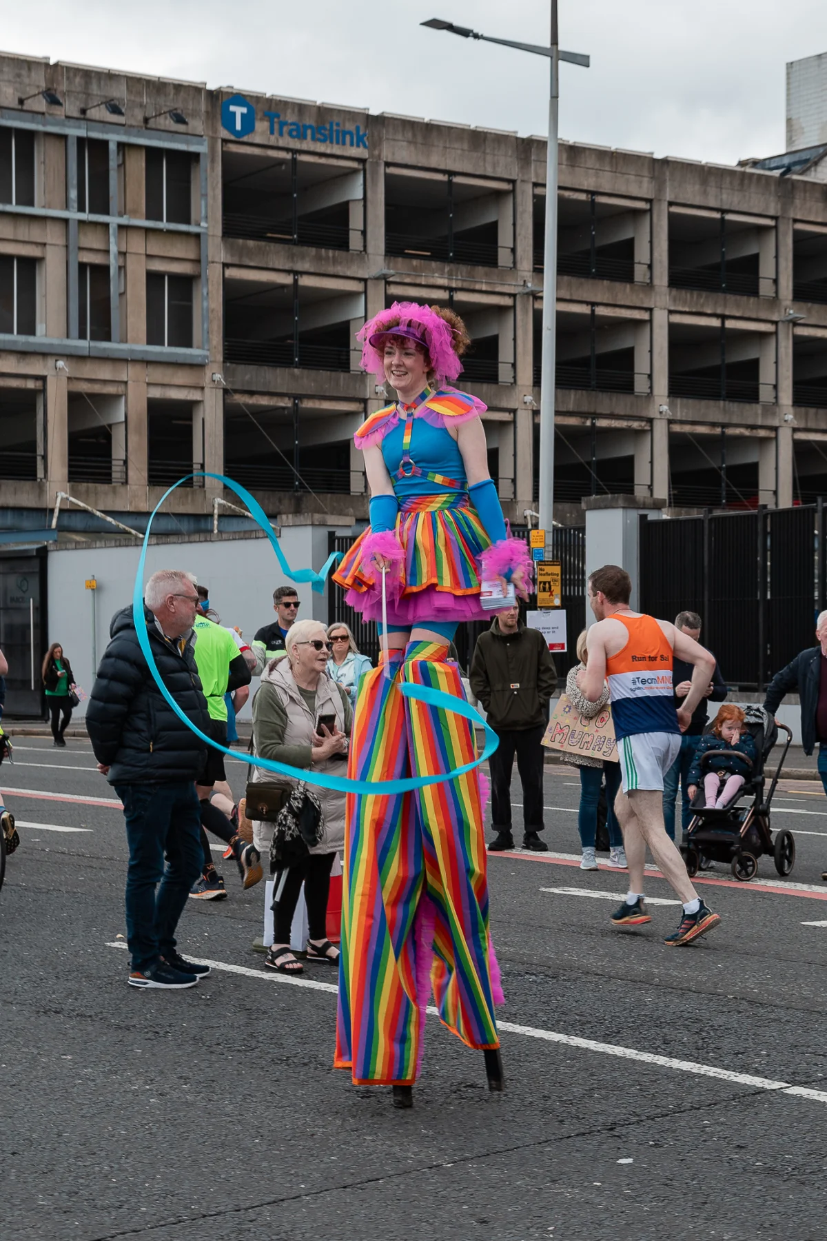 A performer on stilts in a colorful rainbow costume and pink wig at an outdoor urban event.