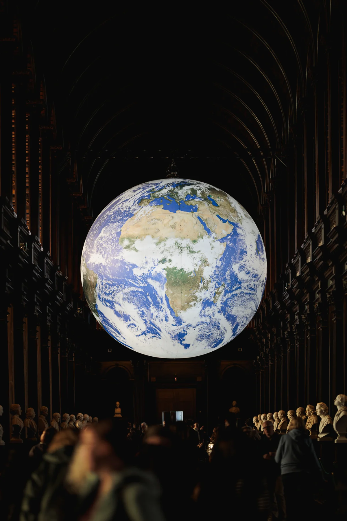 A giant glowing Earth floats in a dark gothic hall filled with statues and blurred silhouettes of people.