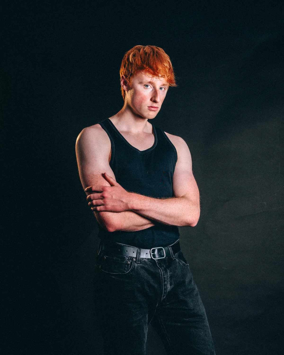 A young man with red hair in a black tank top and jeans posing with crossed arms against a dark studio background.