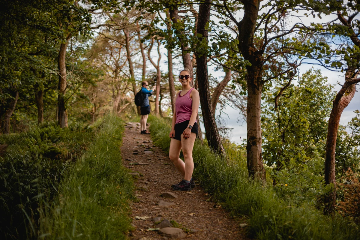 A smiling woman in a pink tank top and black shorts standing on a wooded hiking trail.