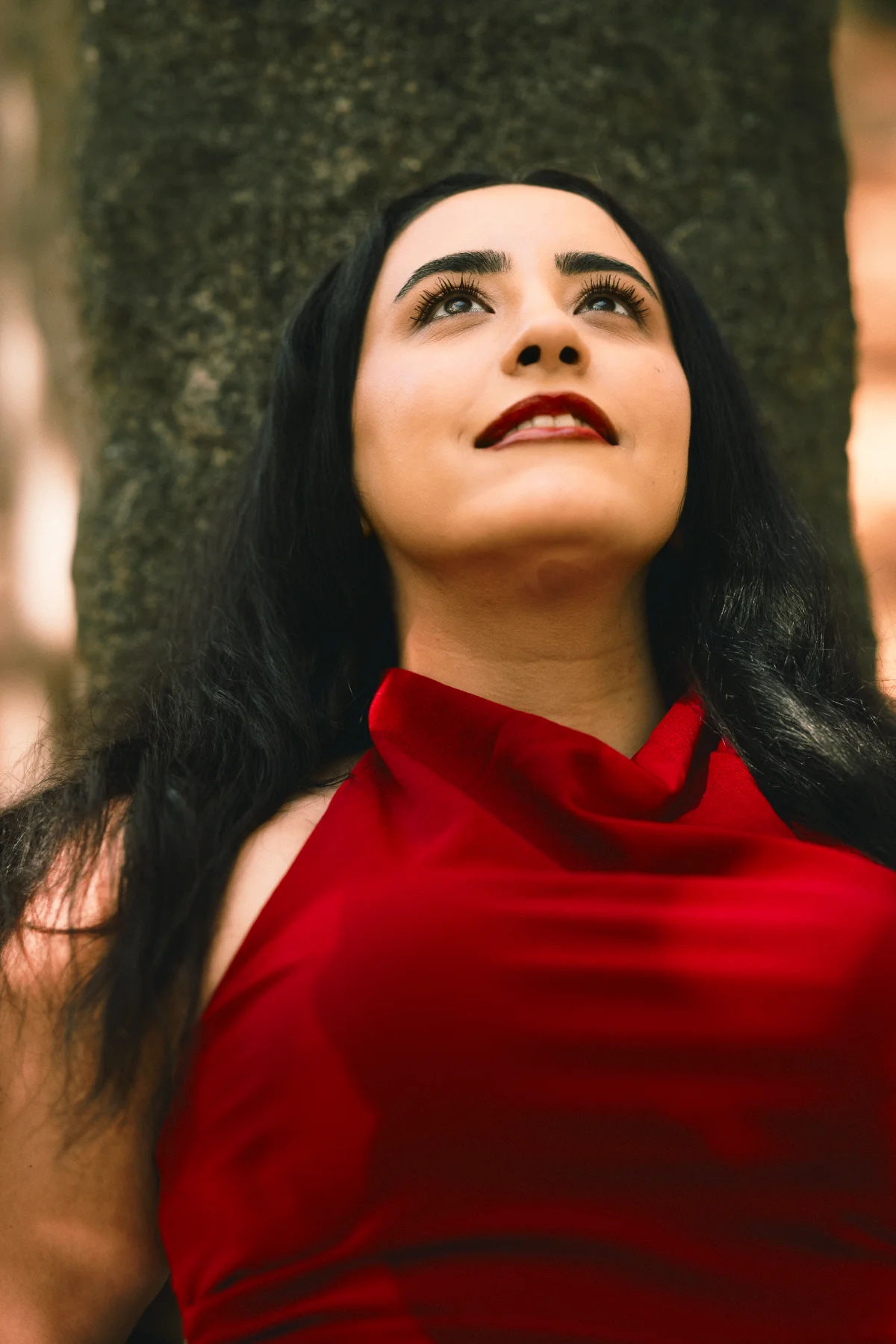 Low-angle portrait of a woman with long black hair and red lipstick wearing a red dress against a tree trunk.