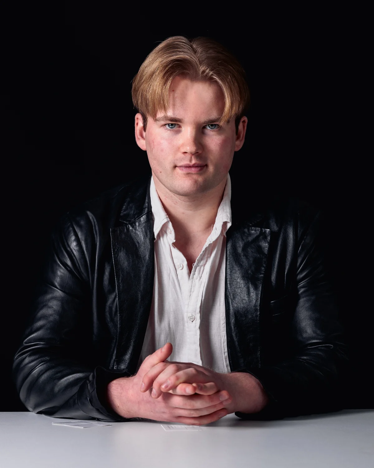 Studio portrait of a young man in a black leather jacket and white shirt against a dark background.