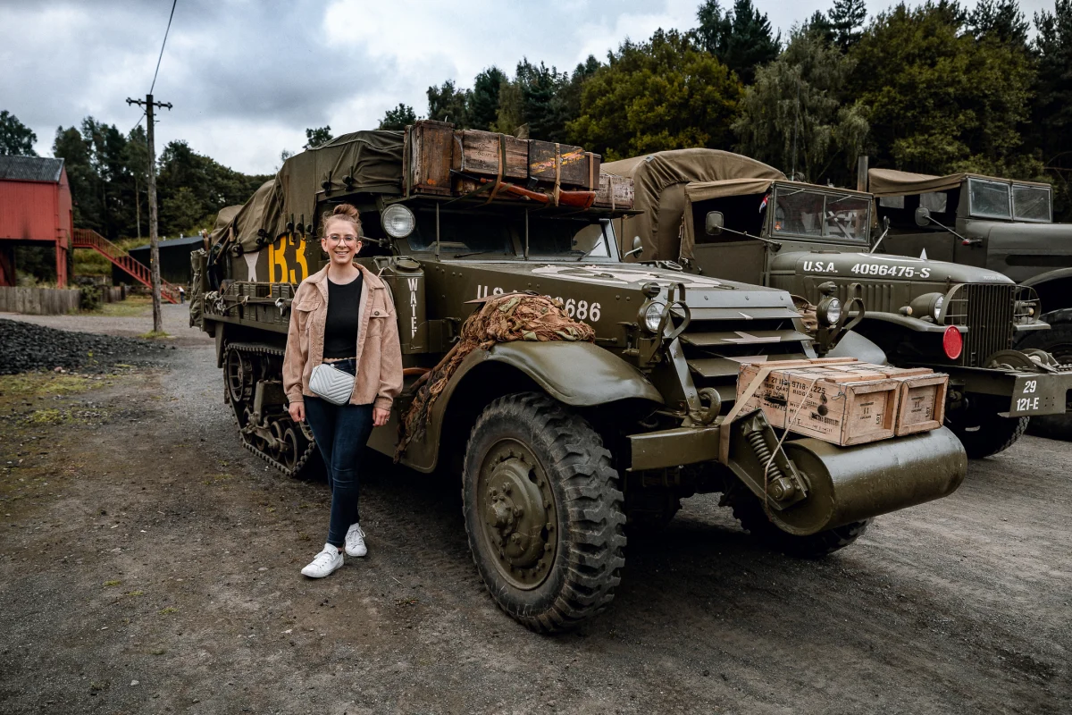 A smiling woman standing next to vintage military vehicles in an outdoor setting.