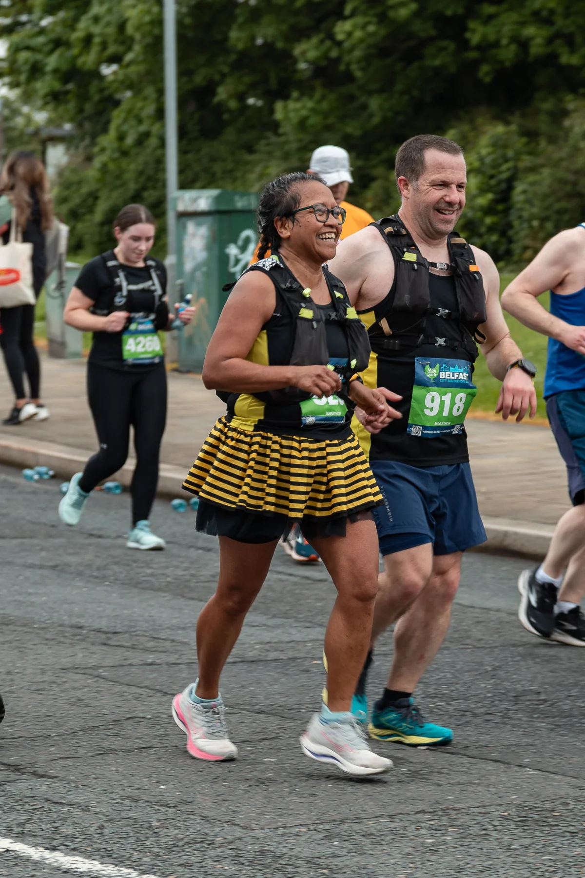 A smiling woman in a yellow and black striped skirt running a race outdoors with other participants.