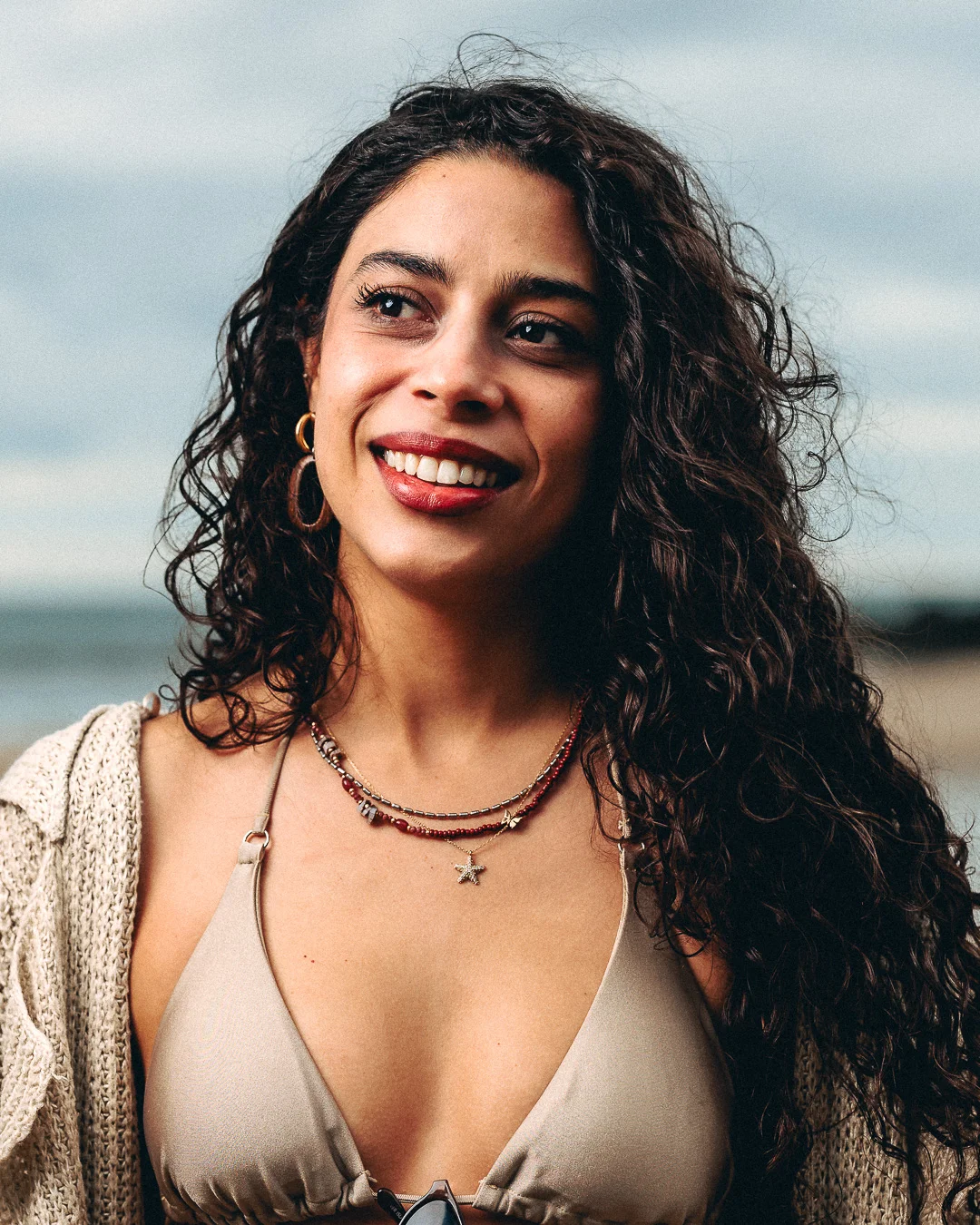 Close-up portrait of a smiling woman with curly dark hair wearing a beige top and necklaces outdoors.