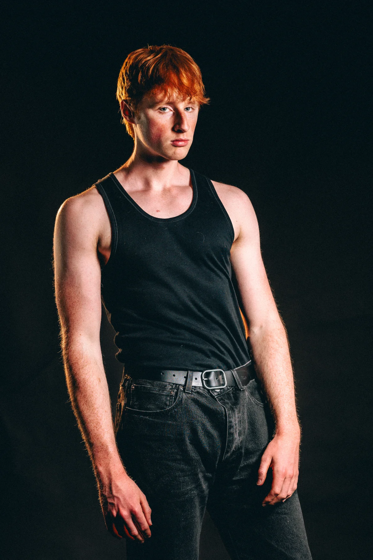 A studio portrait of a young man with red hair wearing a black tank top and jeans against a dark background.