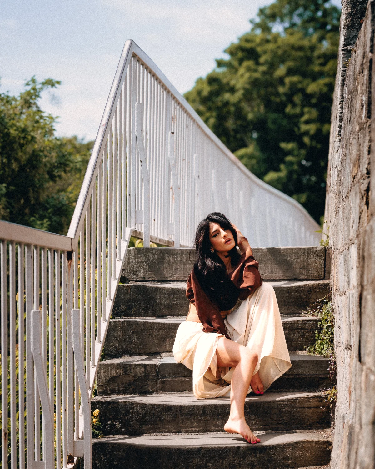 Woman with long dark hair posing on stone stairs beside a white railing in a natural outdoor setting.