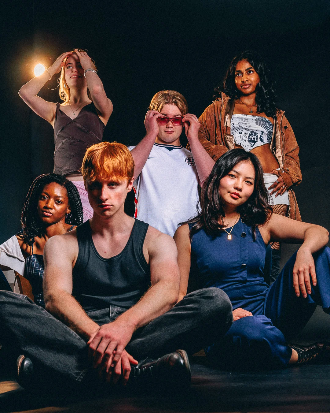 A group of five diverse young adults posing together against a dark backdrop with dramatic studio lighting.