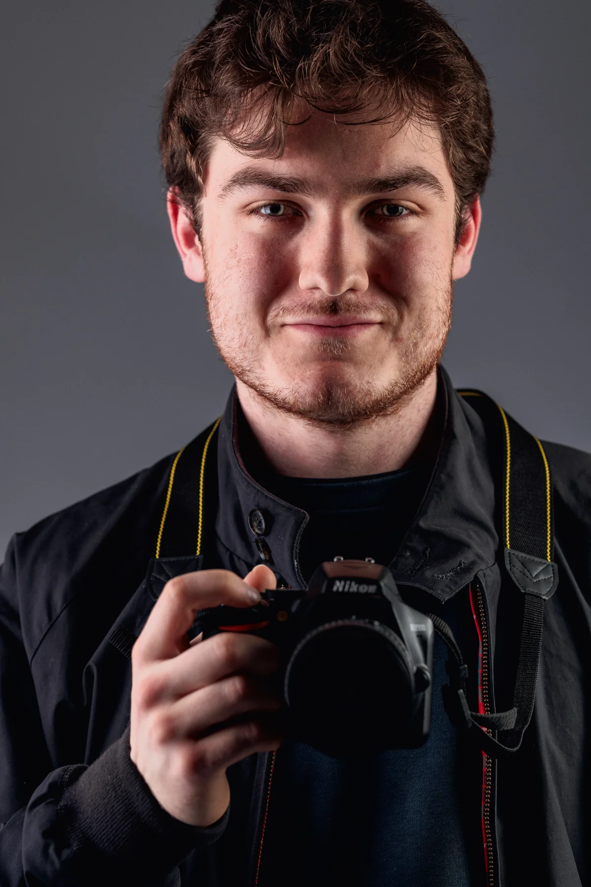 A young man with brown hair and a slight beard holding a Nikon camera against a grey studio background.