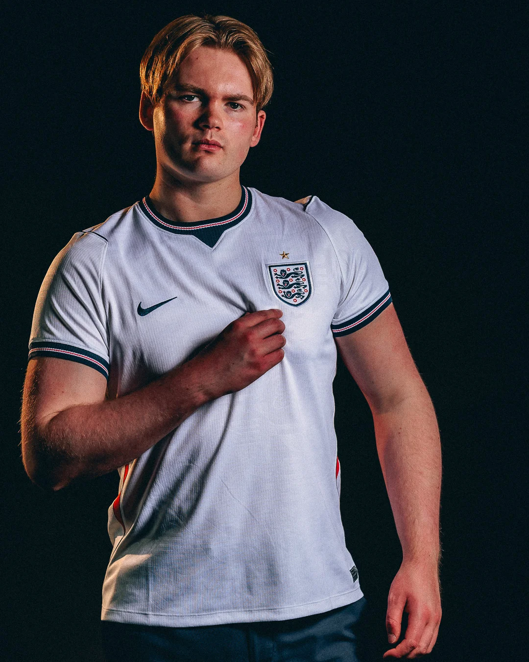 A young man with blonde hair wearing an England national football team jersey against a dark studio background.