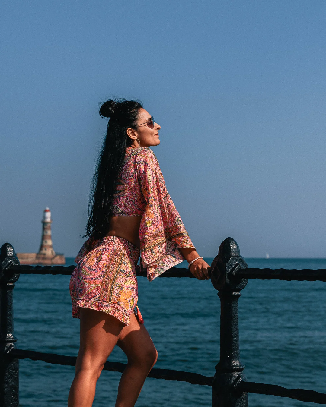 A woman in a colorful patterned two-piece set posing by a seaside railing with a lighthouse in the background.