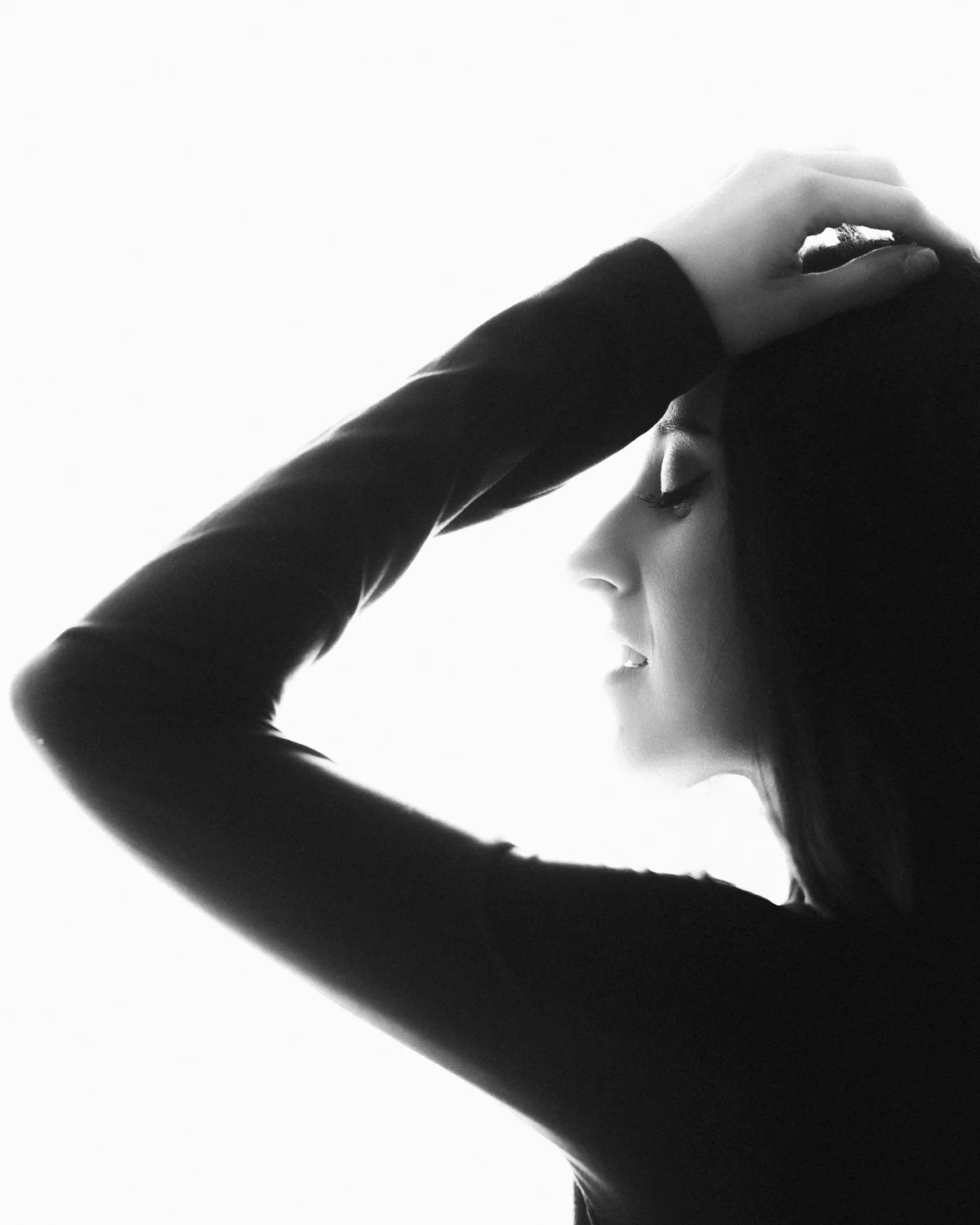 Black and white silhouette profile of a woman with her arm raised to her head against a bright white background.