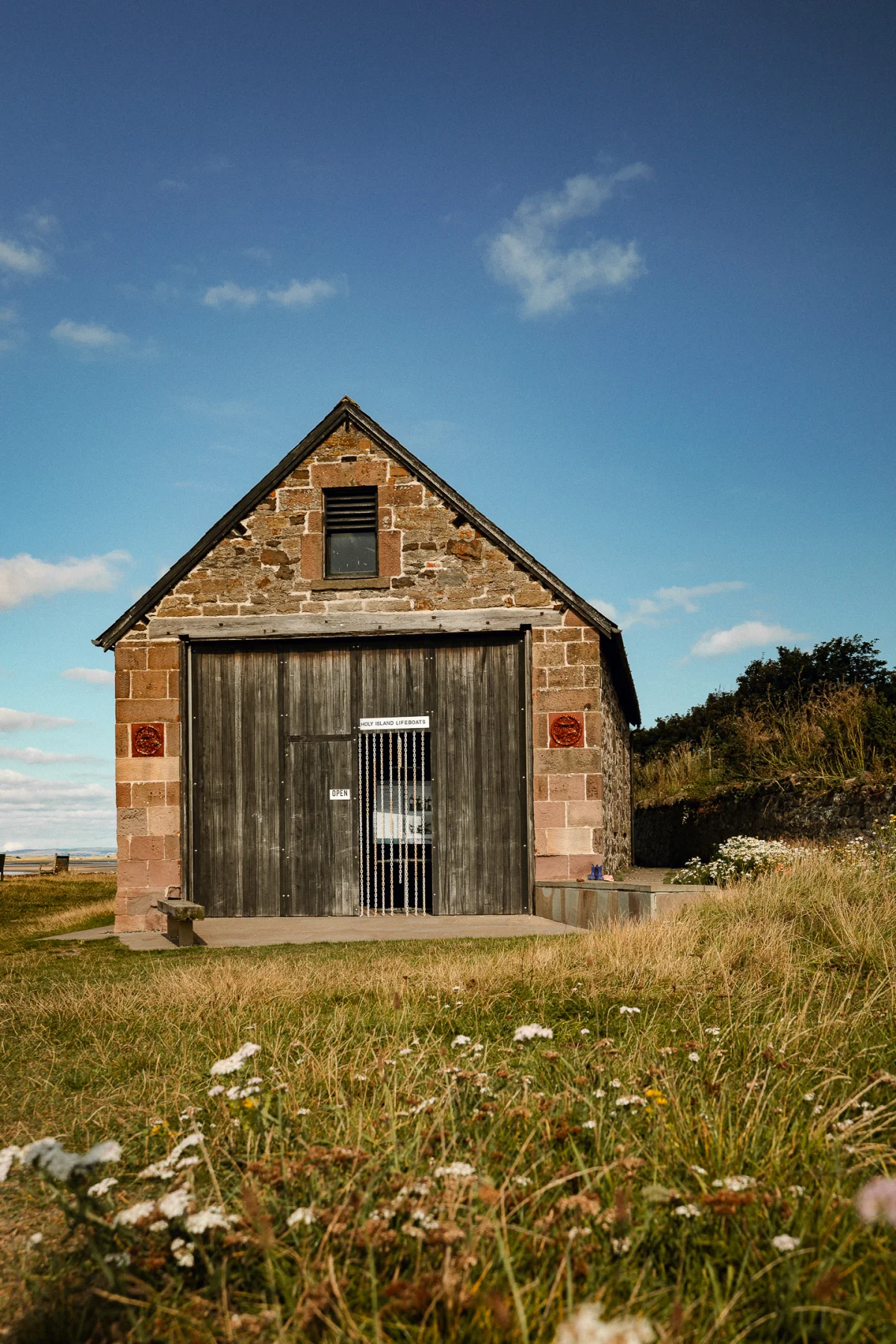 A rustic stone barn with large wooden doors in a grassy field under a clear blue sky.