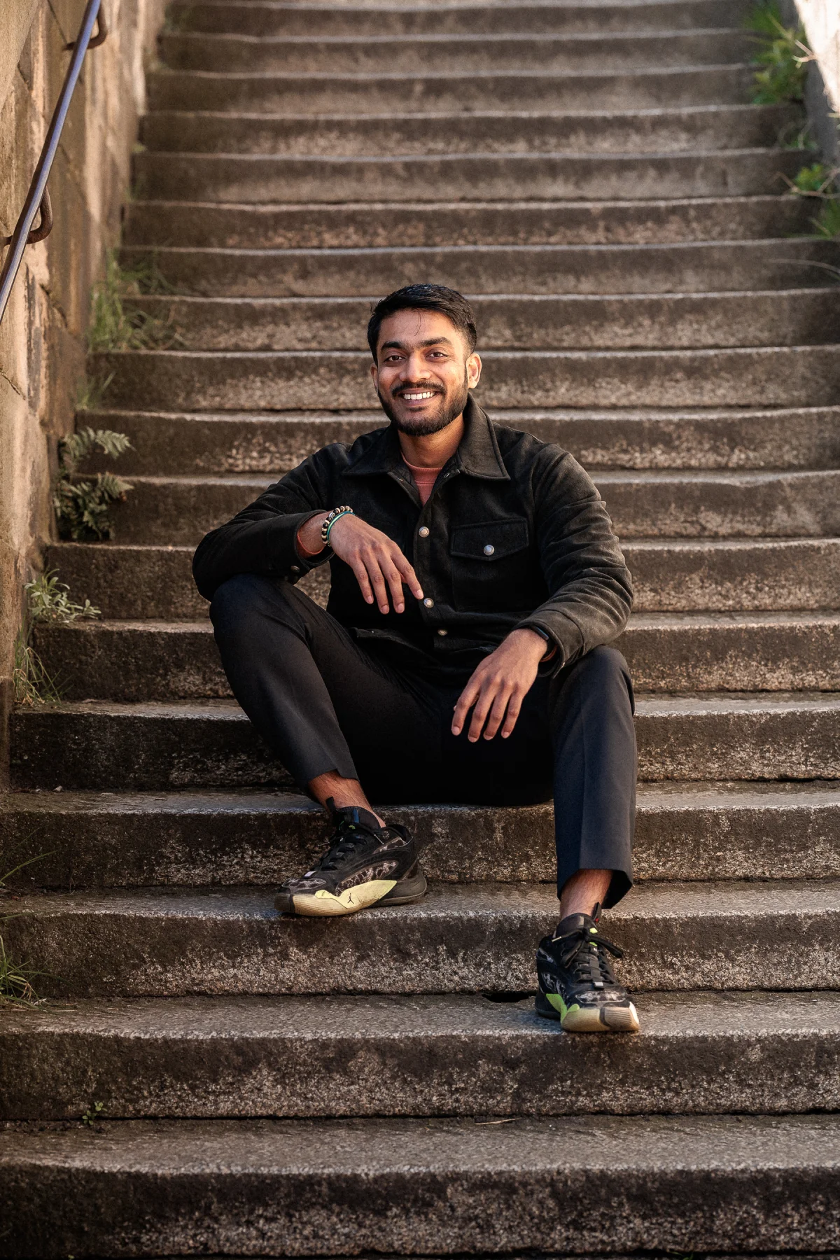 A smiling man with a beard sitting on concrete stairs outdoors wearing all black attire.