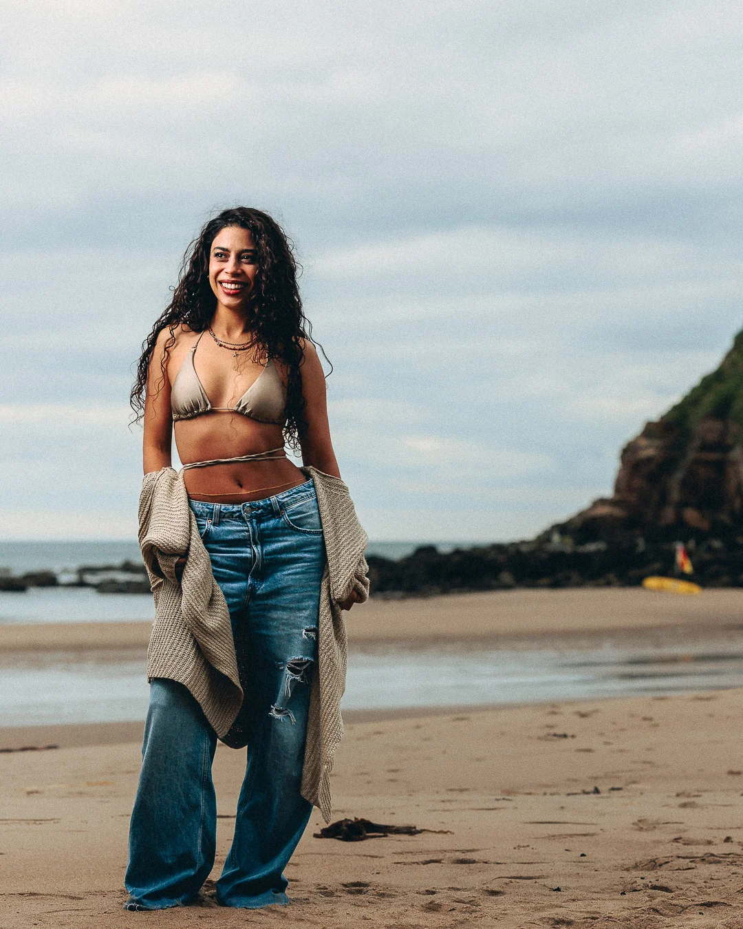 A smiling woman with curly hair wearing a gold bikini top and ripped blue jeans on a sandy beach.