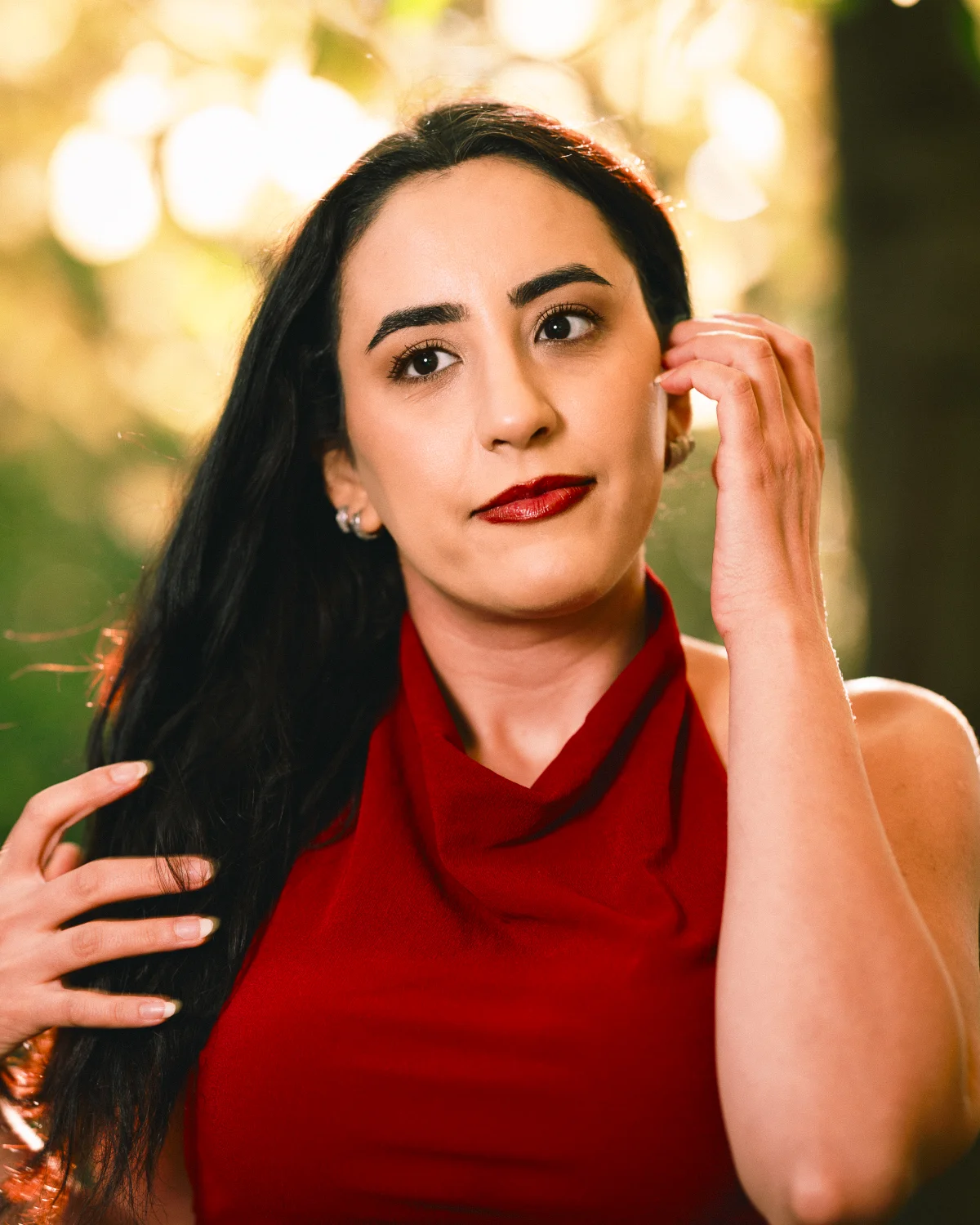 Woman with long dark hair in a red dress posing outdoors against a soft bokeh background.