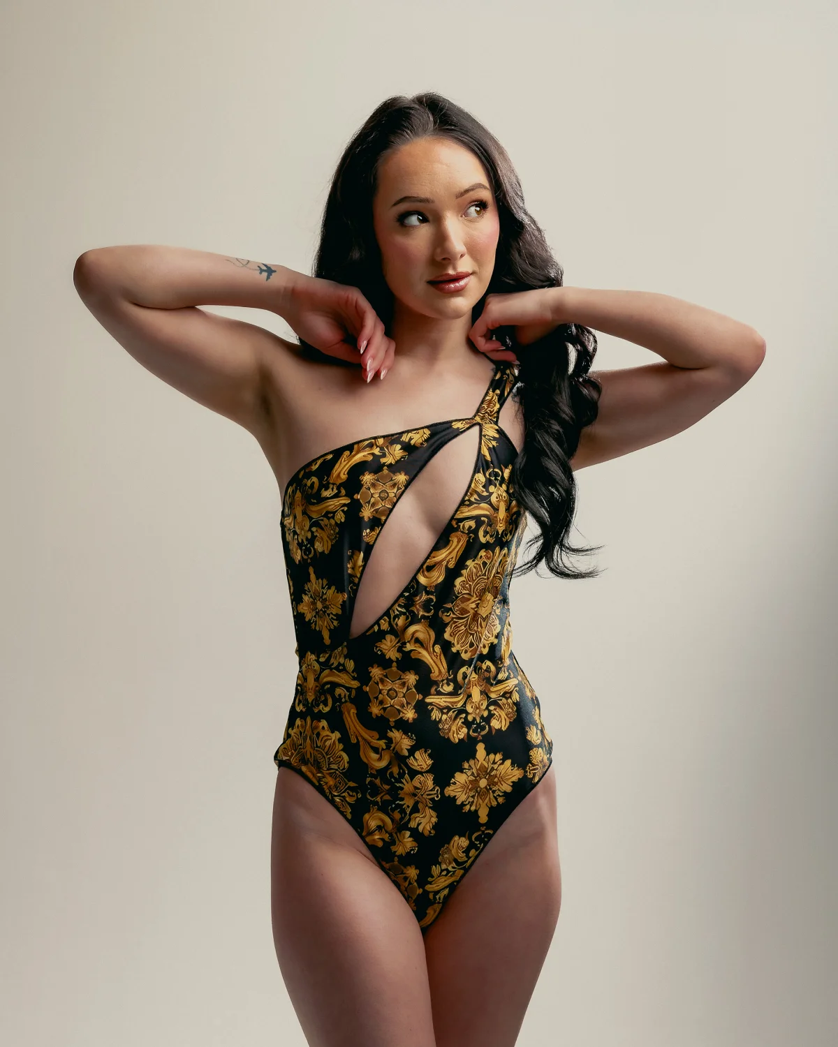 Woman with long dark hair posing in a black and gold patterned one-shoulder swimsuit against a neutral studio background.