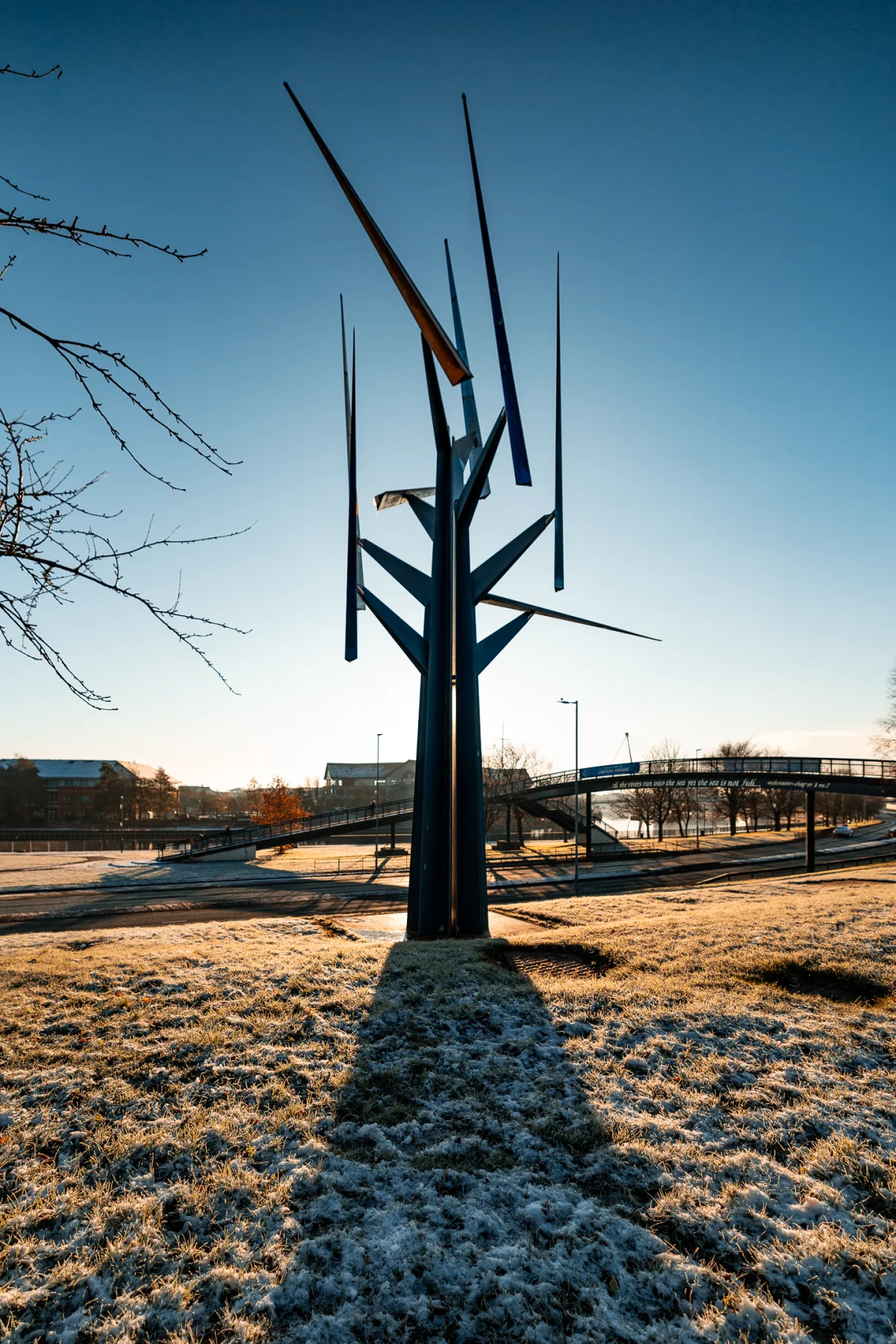 A tall abstract metal sculpture in a frosty field under a clear blue sky with a bridge in the background.