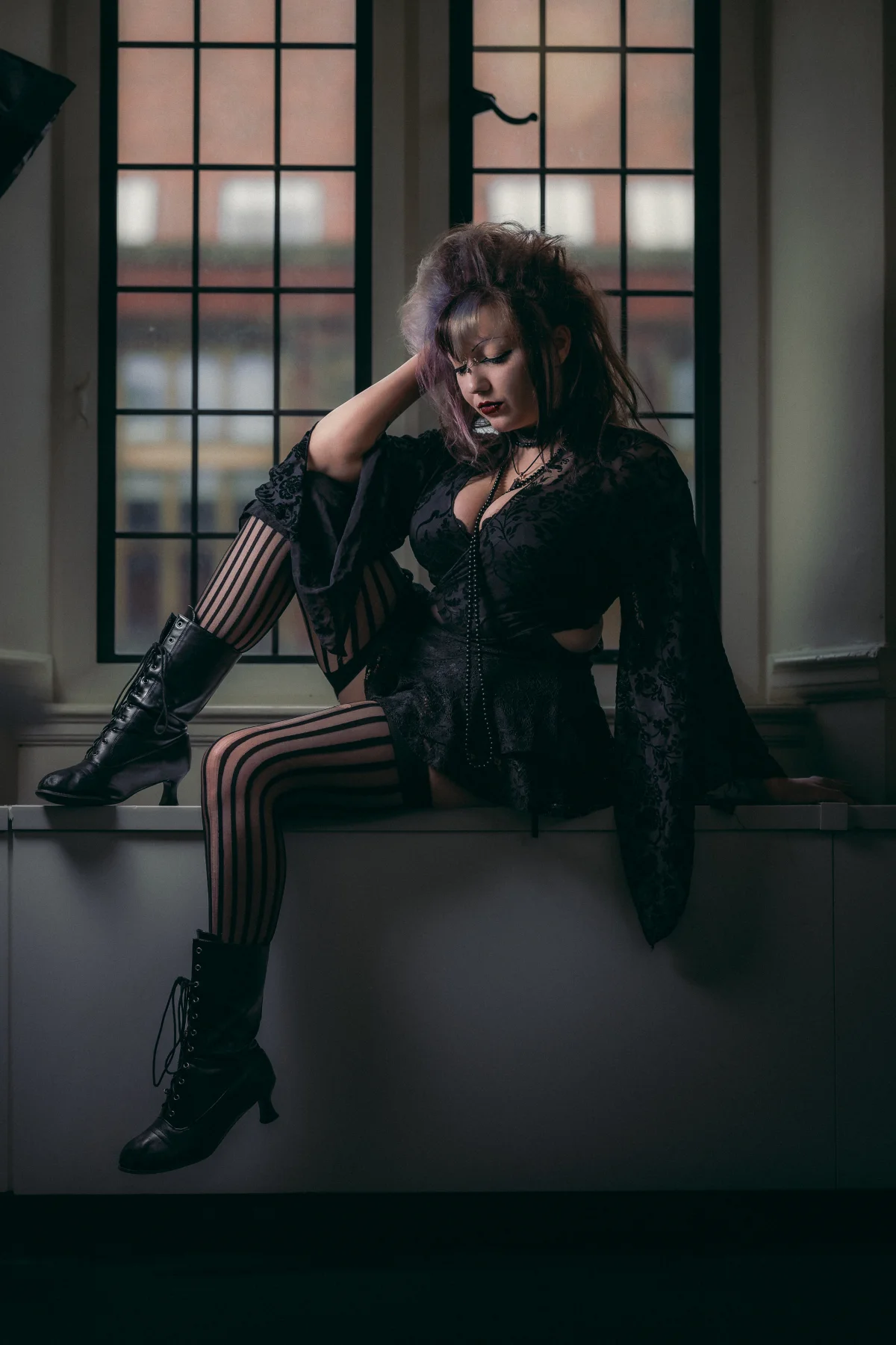 A woman with purple hair in gothic black attire and striped stockings sits moodily by a window.