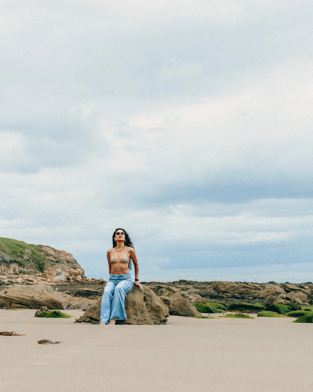 A woman in a bikini top and light blue jeans sits on a rock at a beach under an overcast sky.