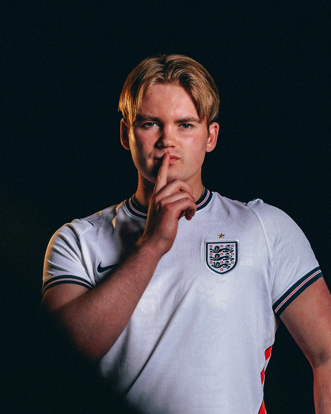 Young man in an England football jersey making a shushing gesture against a dark studio background.