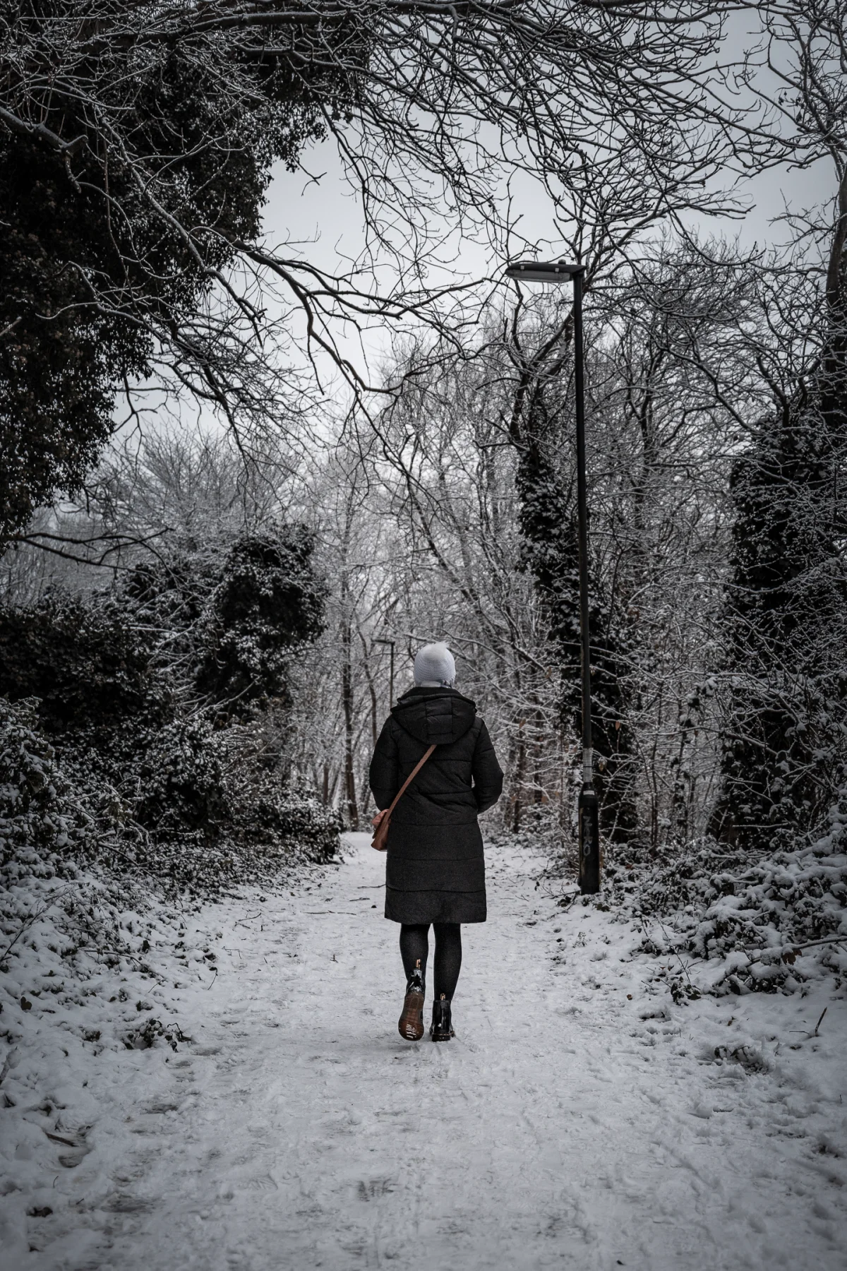 Person in a dark coat and white beanie walking away on a snowy path through frosted trees.