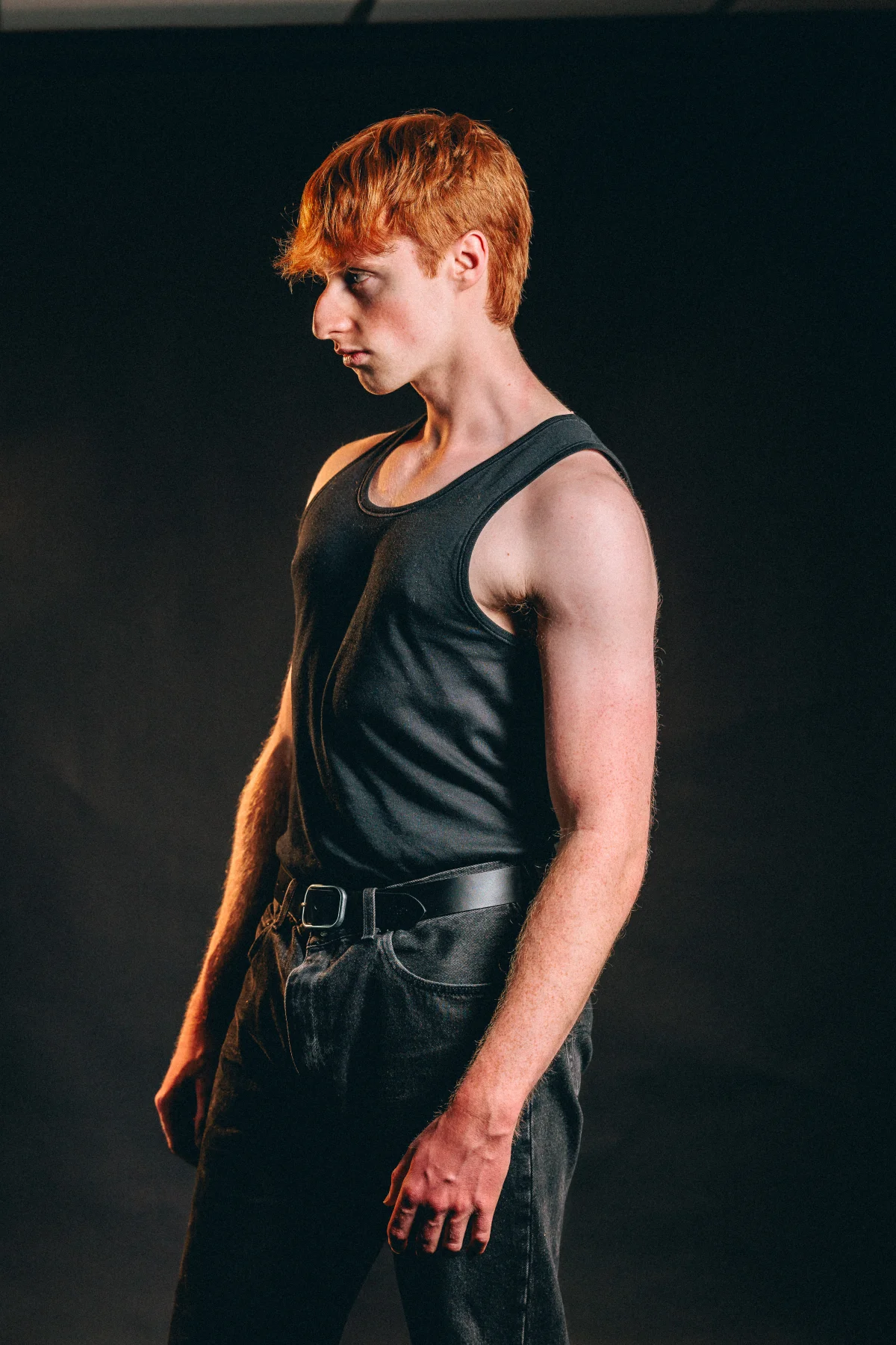 Young man with red hair in a black tank top posing against a dark background.