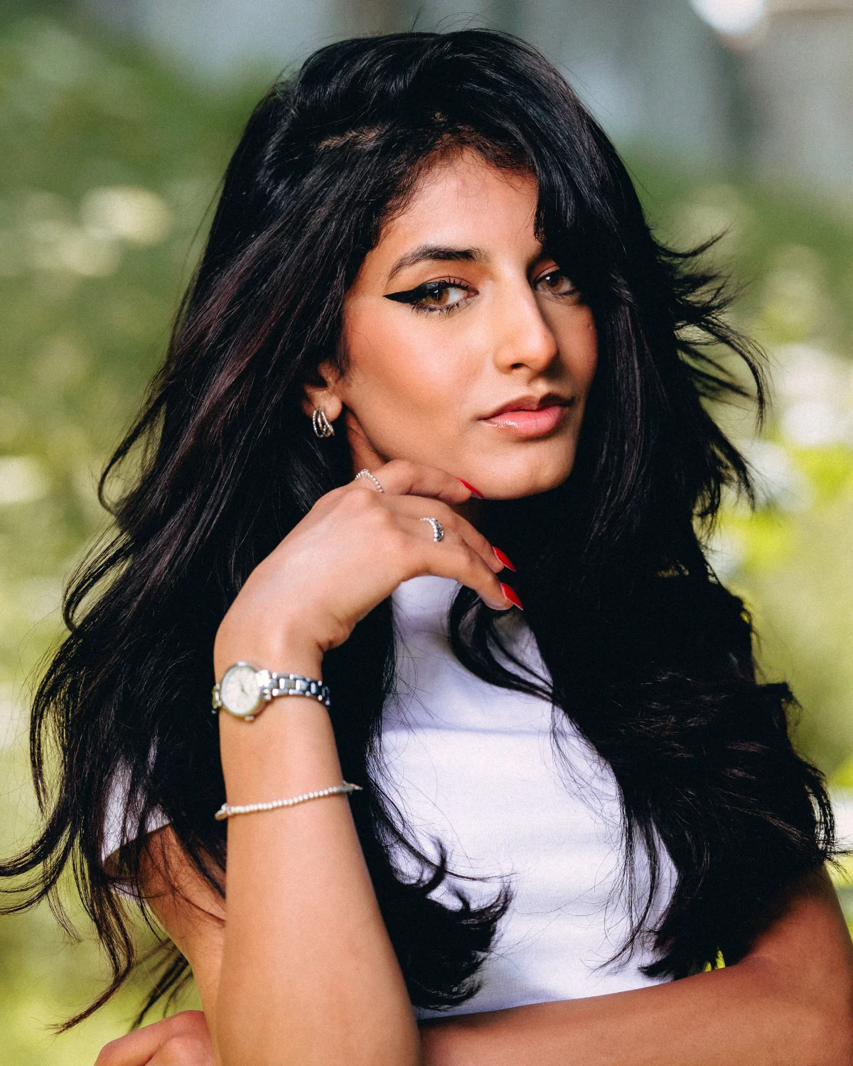Close-up portrait of a woman with long dark hair and bold eyeliner posing outdoors in a white top.