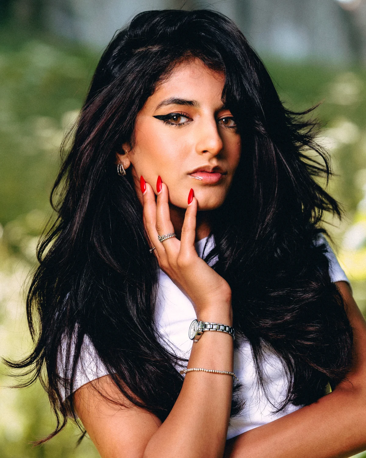 Close-up portrait of a woman with long dark hair and bold eyeliner posing against a blurred green outdoor background.
