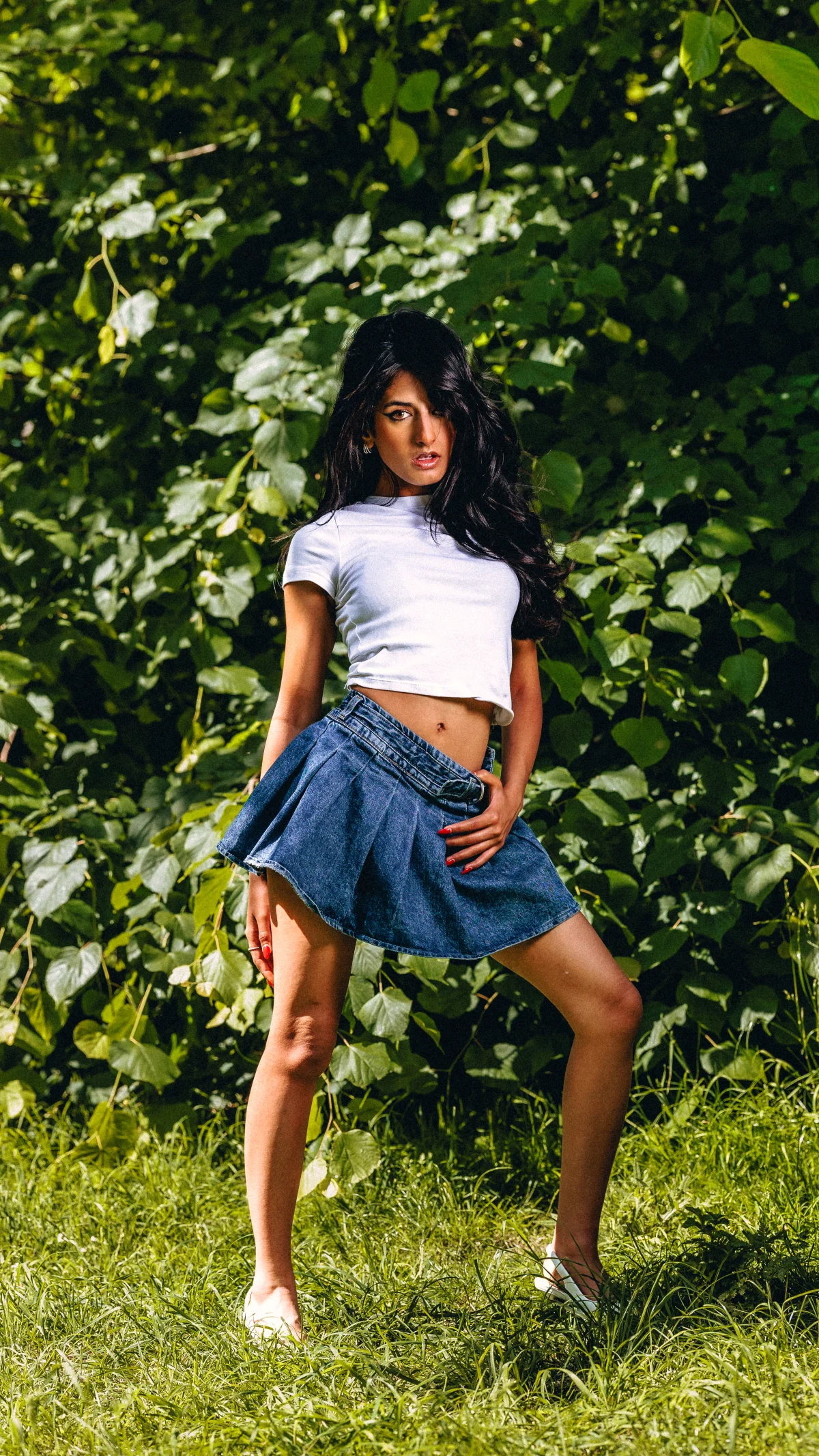 A woman with long dark hair posing in a white crop top and denim skirt against a lush green foliage background.