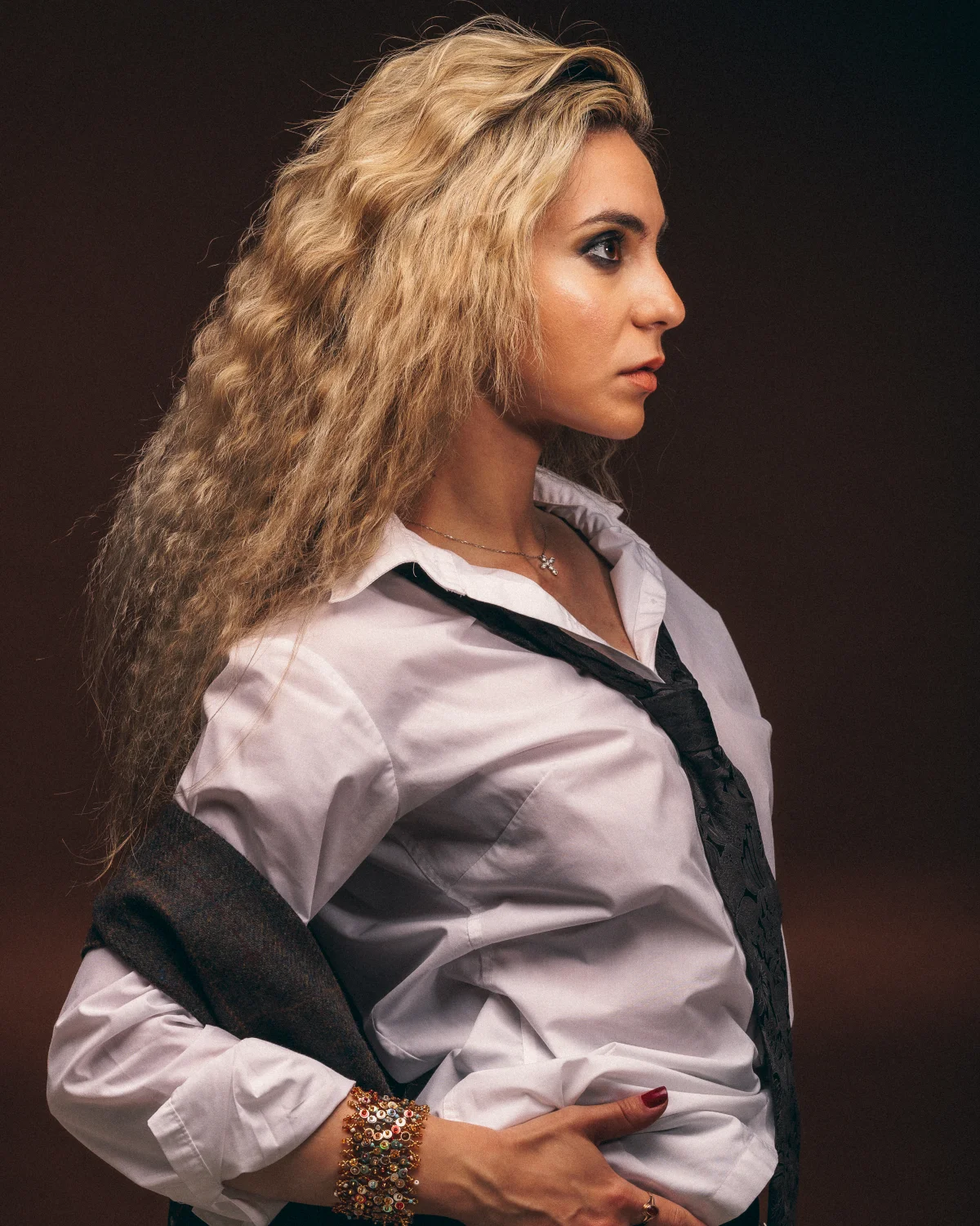 A woman with long blonde wavy hair in a white shirt and loose black tie against a dark brown background.