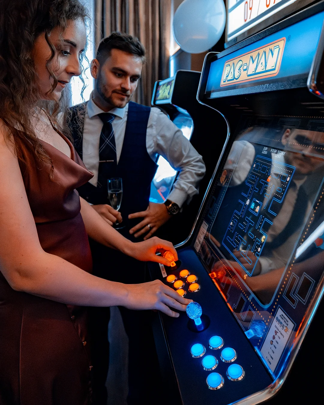 A woman in a red dress and a man in formal attire play Pac-Man at an arcade.