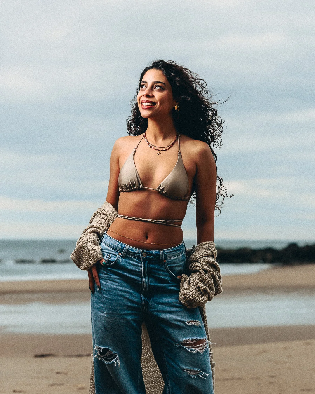 A smiling woman with curly hair wearing a gold bikini top and ripped jeans on a sandy beach.