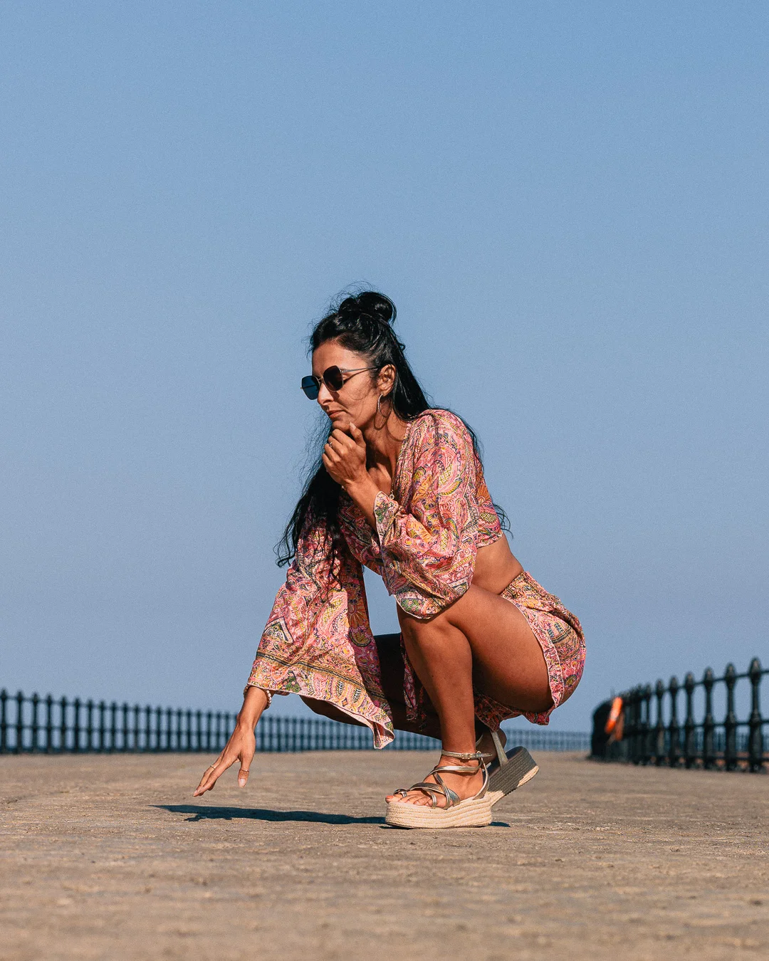 Woman in a colorful patterned two-piece set posing in a crouched position on a paved coastal area under a blue sky.