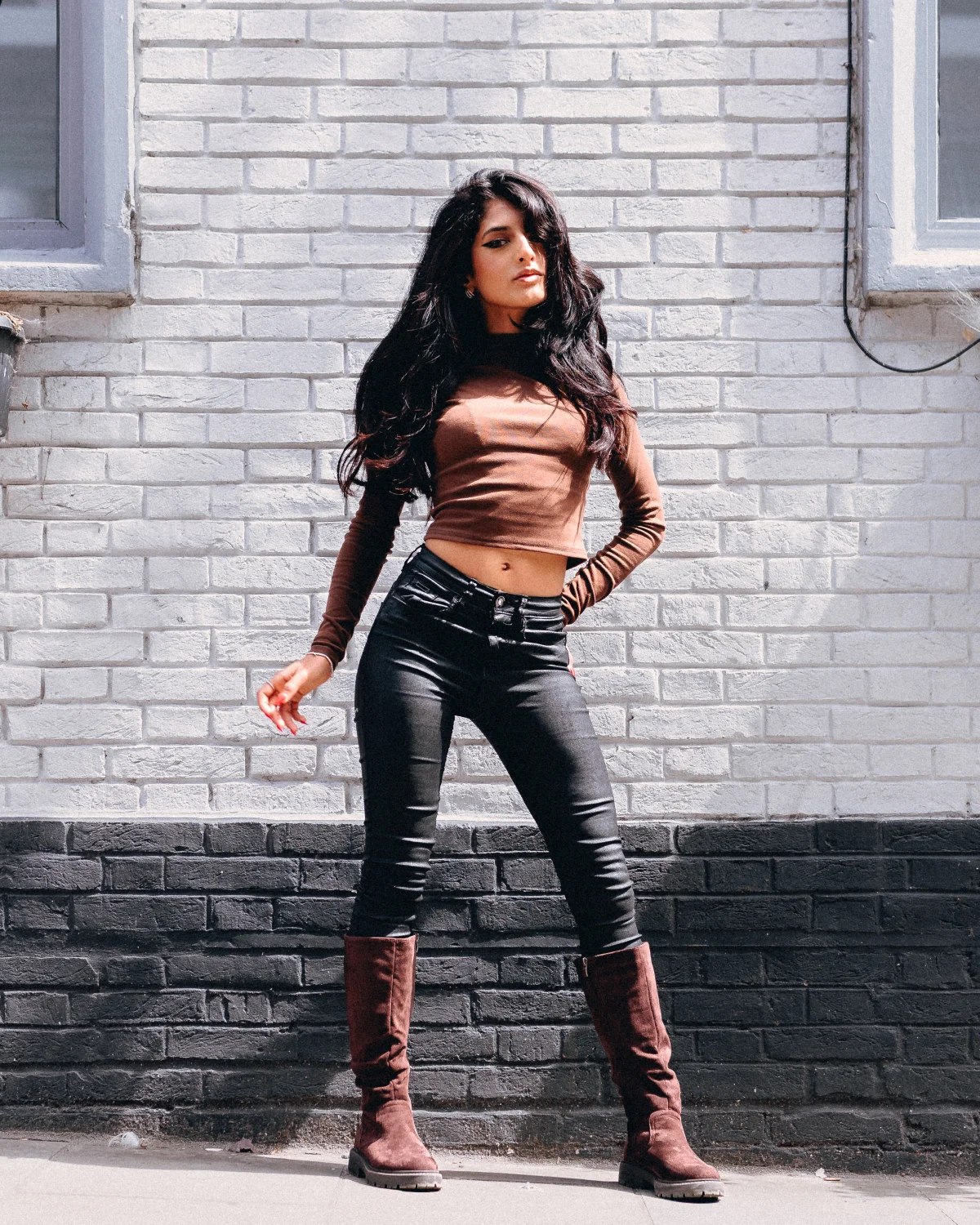 Full-body shot of a woman in a brown crop top and black jeans posing against a white brick wall.