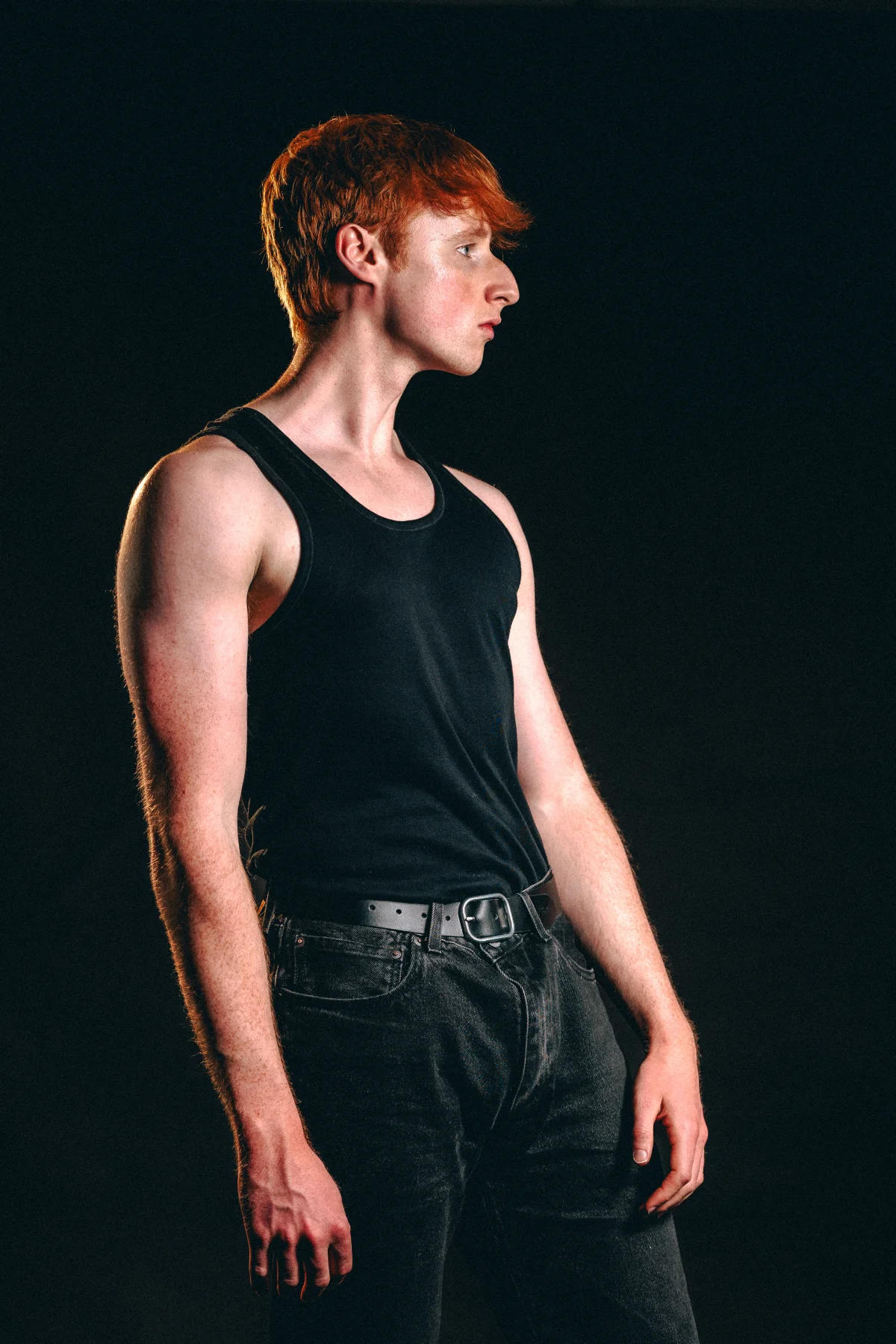 Profile view of a young man with red hair wearing a black tank top and dark jeans against a black background.